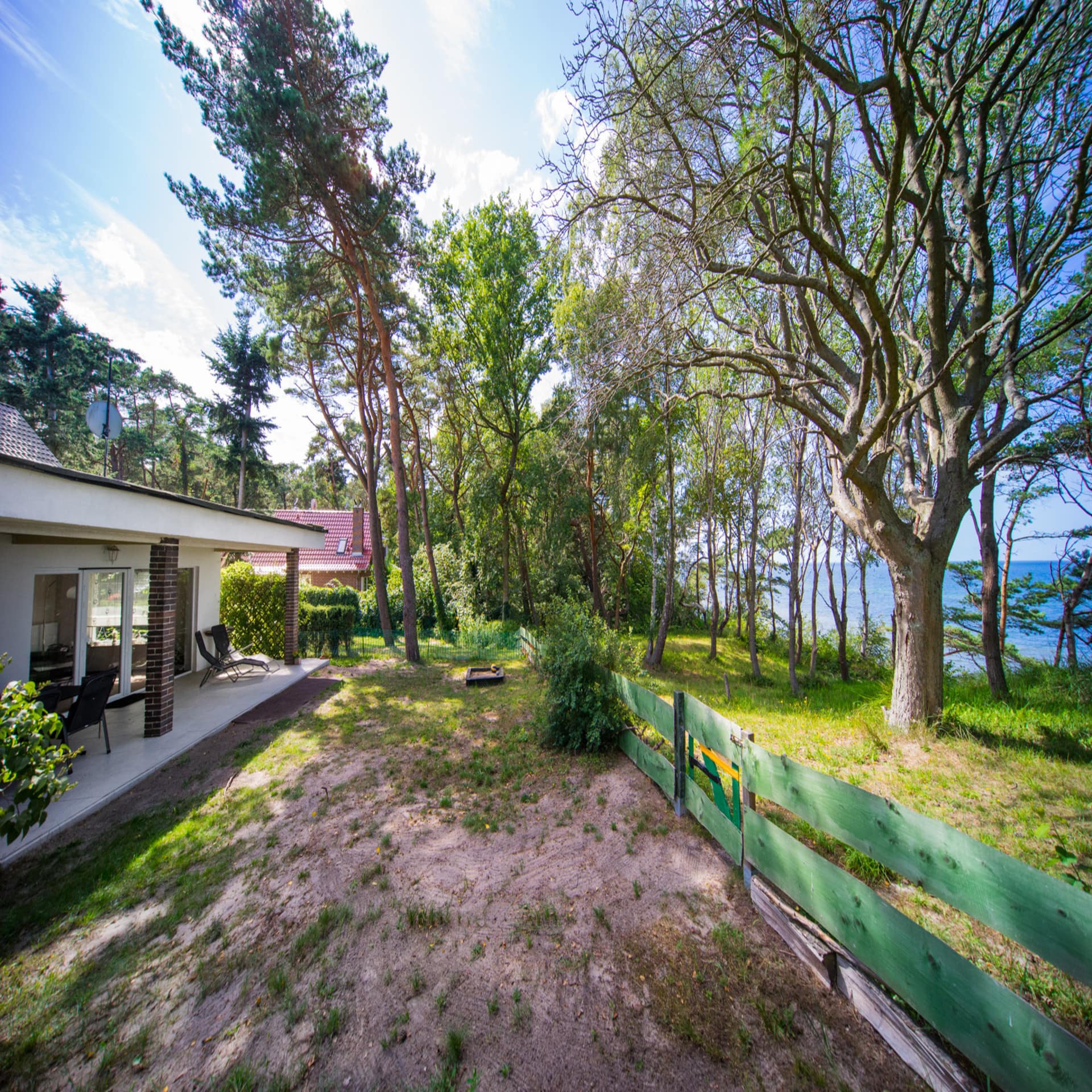 Bungalow mit Garten und grünem Gartenzaun mit Blick durch Bäume auf das Wasser.