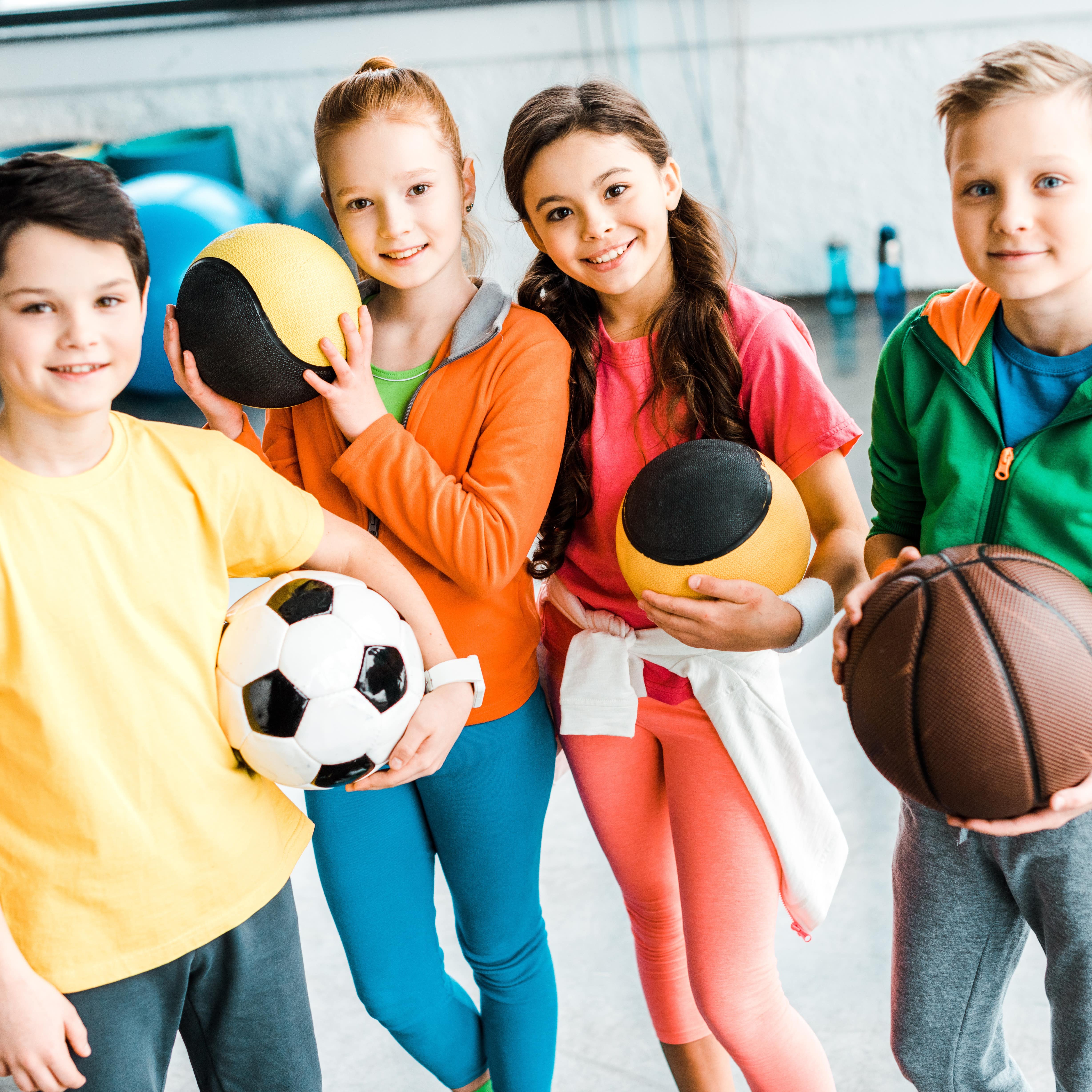 In einer Sporthalle: 4 Kinder in Sportbekleidung mit Bällen in der Hand.