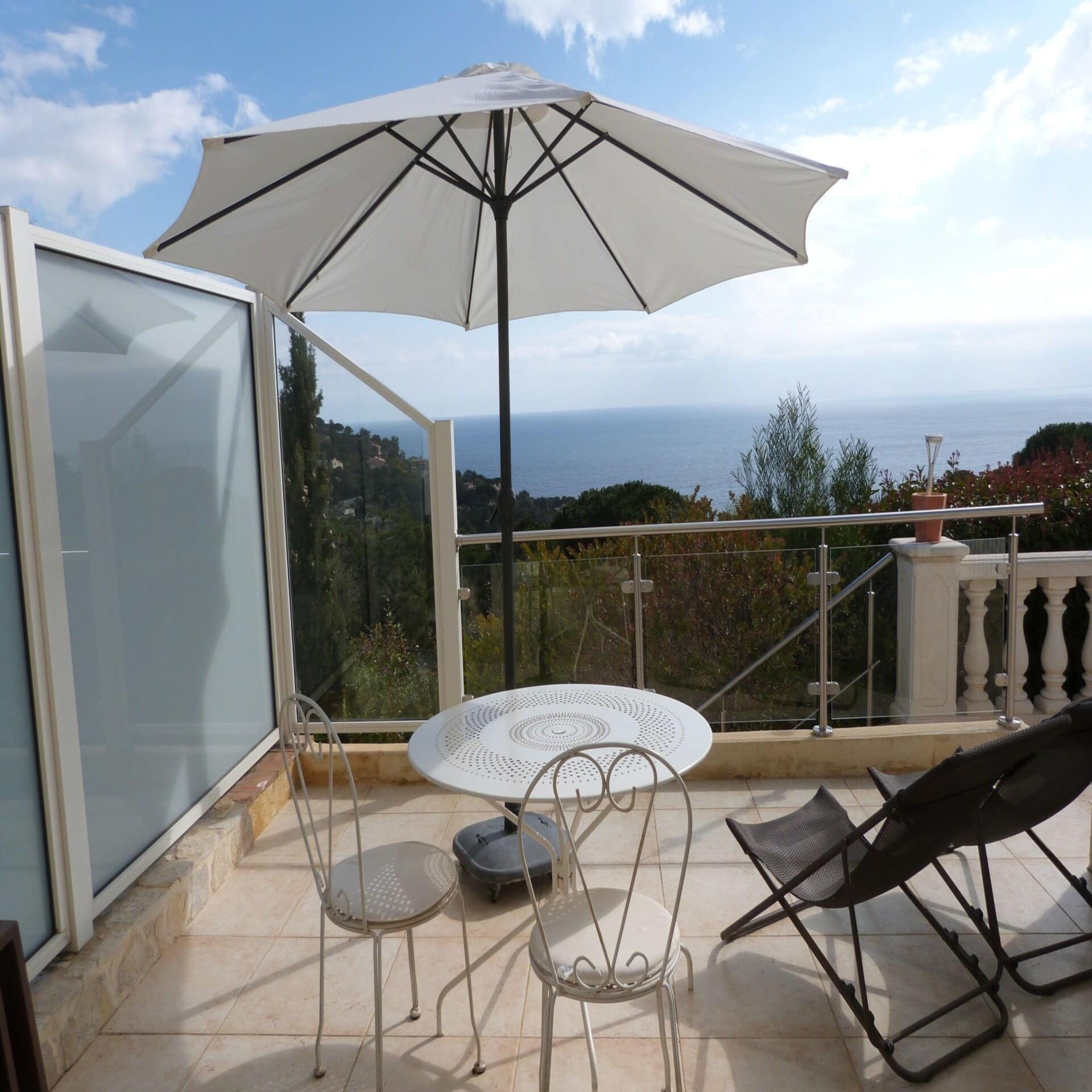 Terrasse mit Tisch, 2 Stühlen, 2 Liegestühlen und Sonnenschirm sowie Blick über Bäume auf das Mittelmeer.