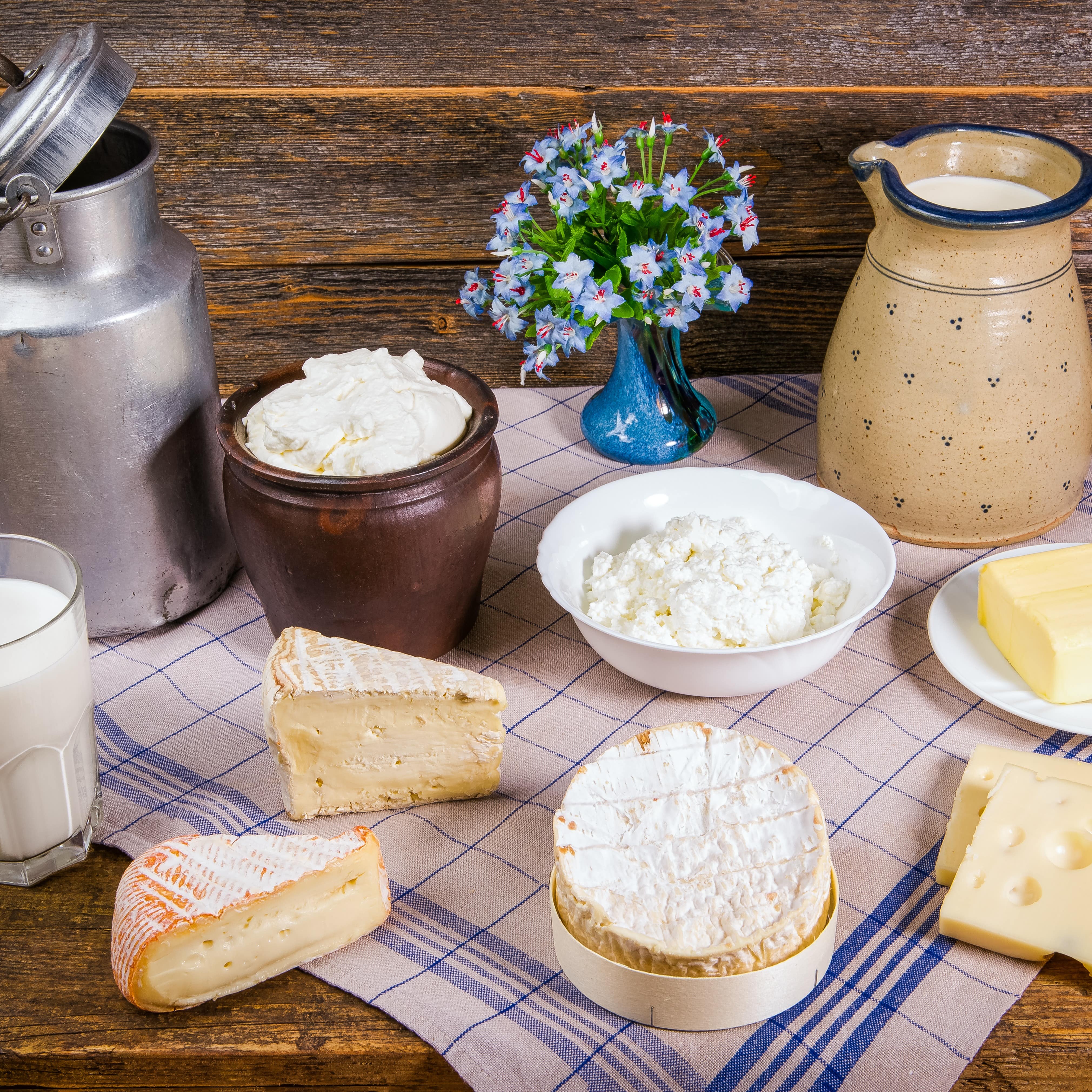 Auf einem Holztisch mit Küchentuch stehen verschiedene Käsesorten, eine Milchkanne, ein Milchkrug und ein Glas Milch. 