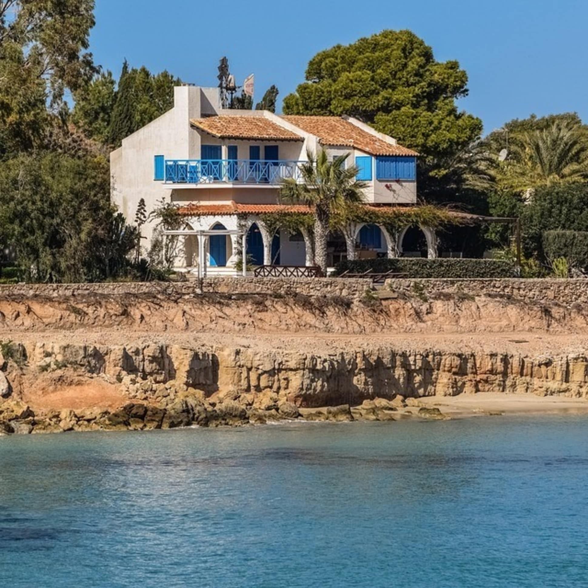Villa mit blauem Balkon und Rundtorbögen am Meer mit Palmen im Hintergrund