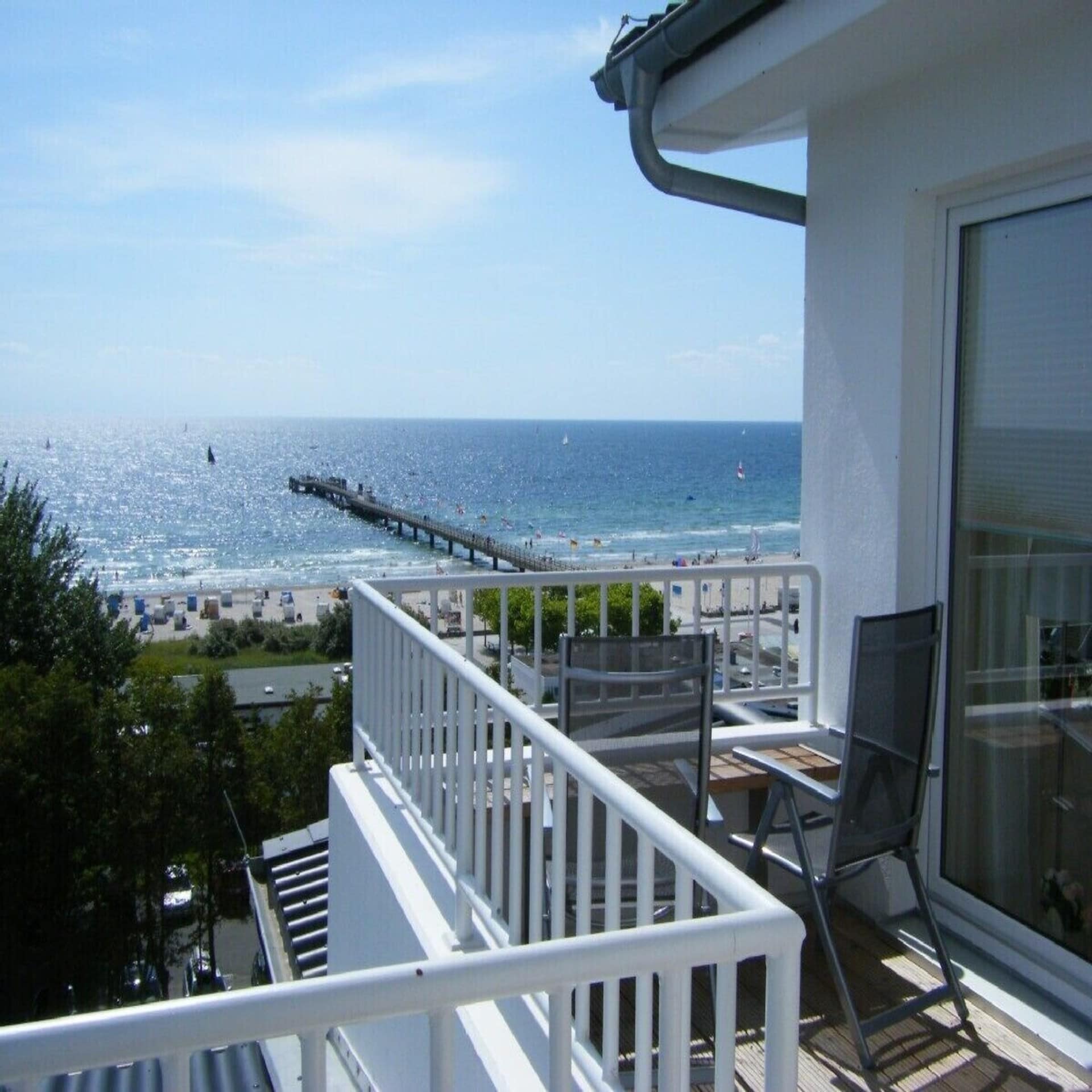 Sonniger Balkon mit Stühlen und Blick auf die Ostsee und eine Seebrücke am Strand.