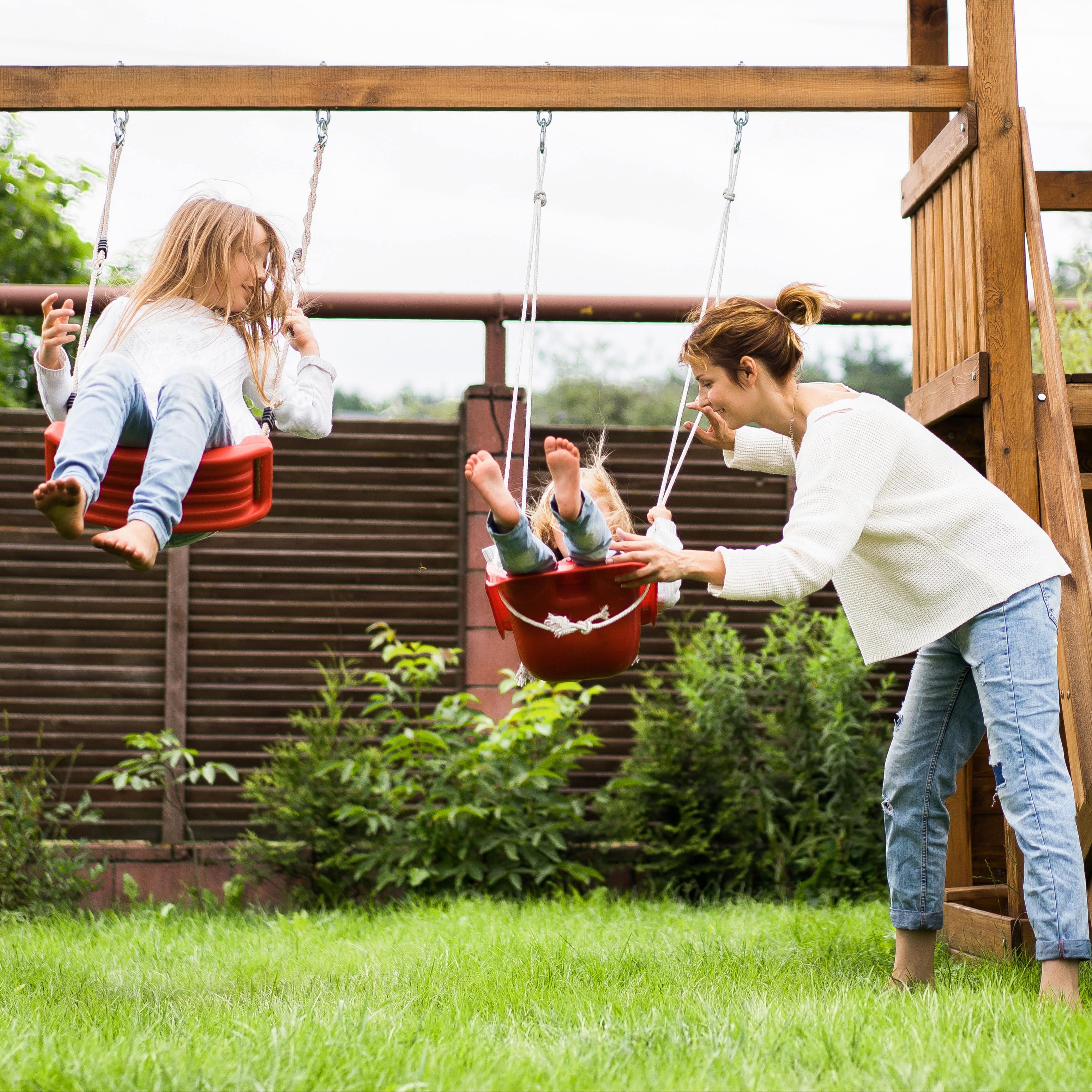 Mutter schaukelt 2 Mädchen auf einem Klettergerüst mit Schaukel in einem Garten.