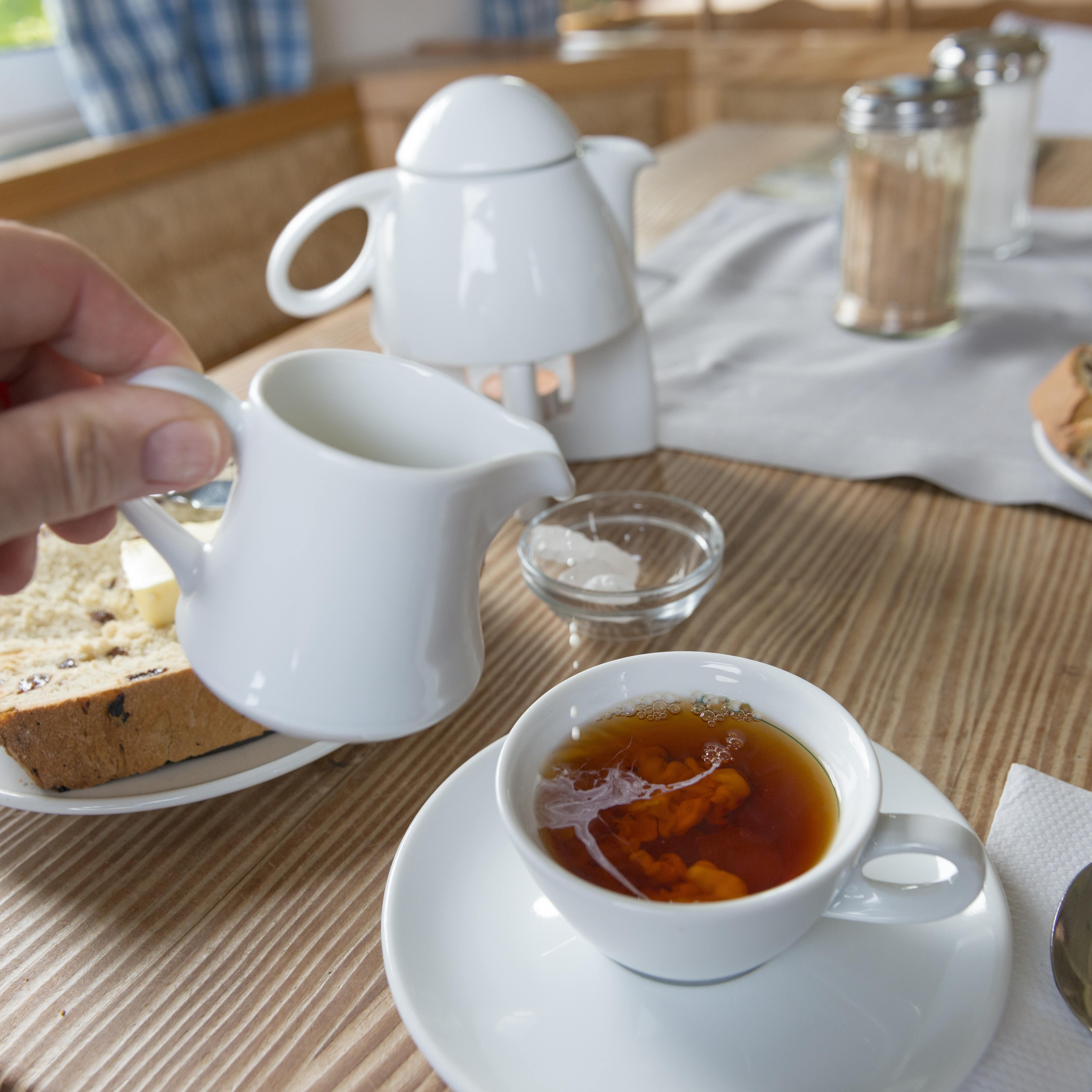  Teestunde: Eine Hand gießt Sahne in den Tee, rund herum Kandiszucker und Rosinenstuten mit Butter.