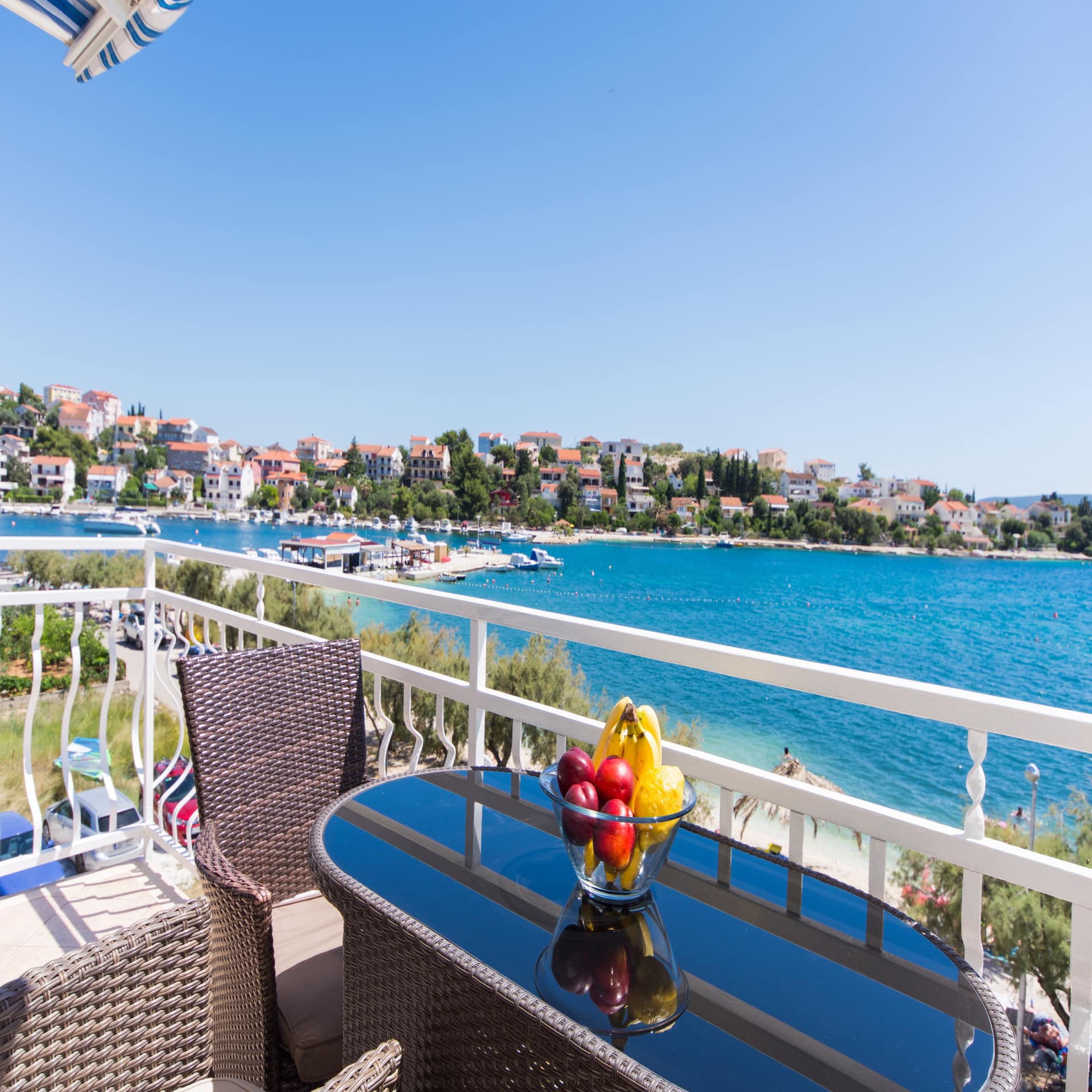 Sonniger Balkon mit Tisch, Stühlen und Obstschale direkt am Meer mit Blick auf das Wasser und den Ort.