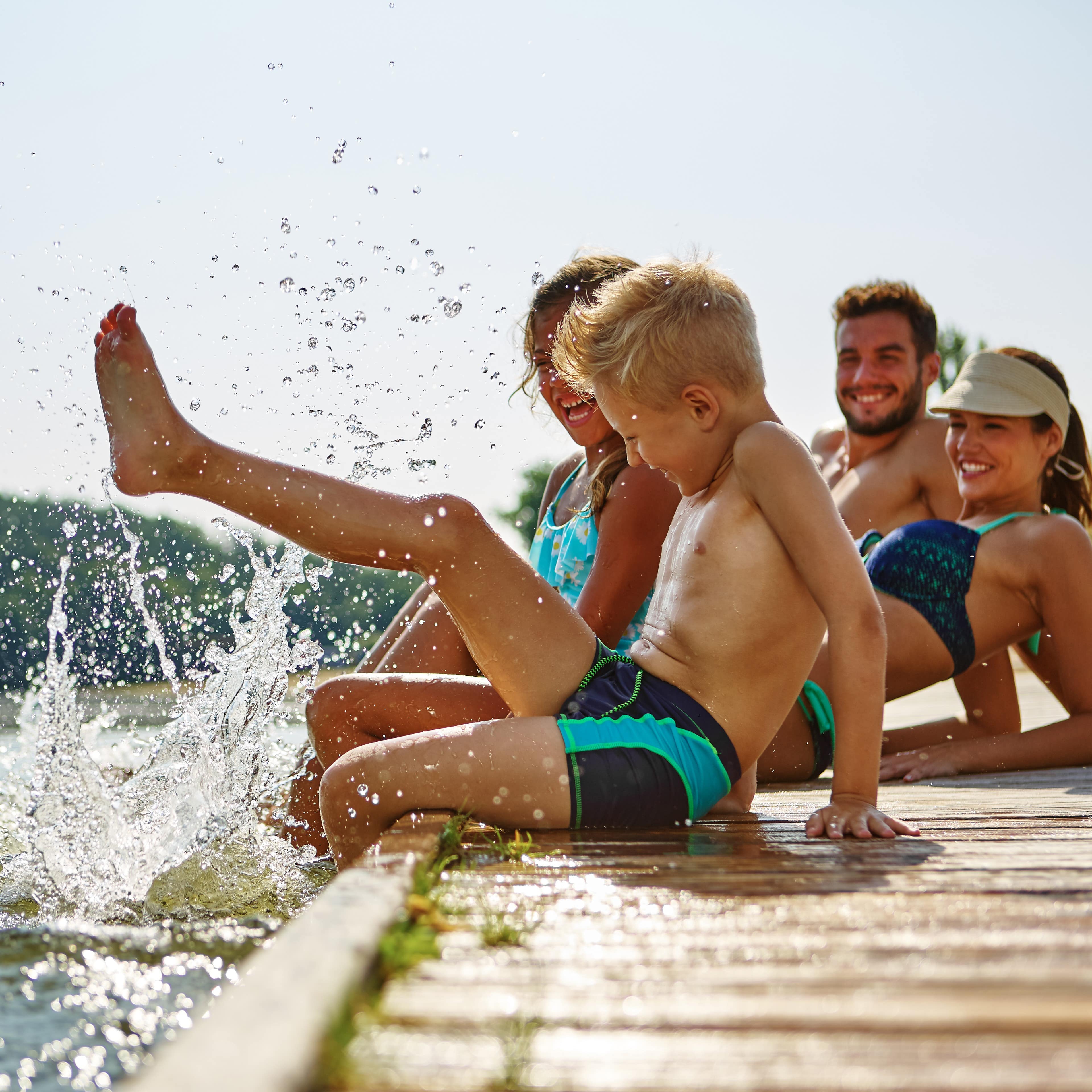 Familie mit 2 Kindern in Badesachen auf einem Holzsteg am See planscht am Wasser.