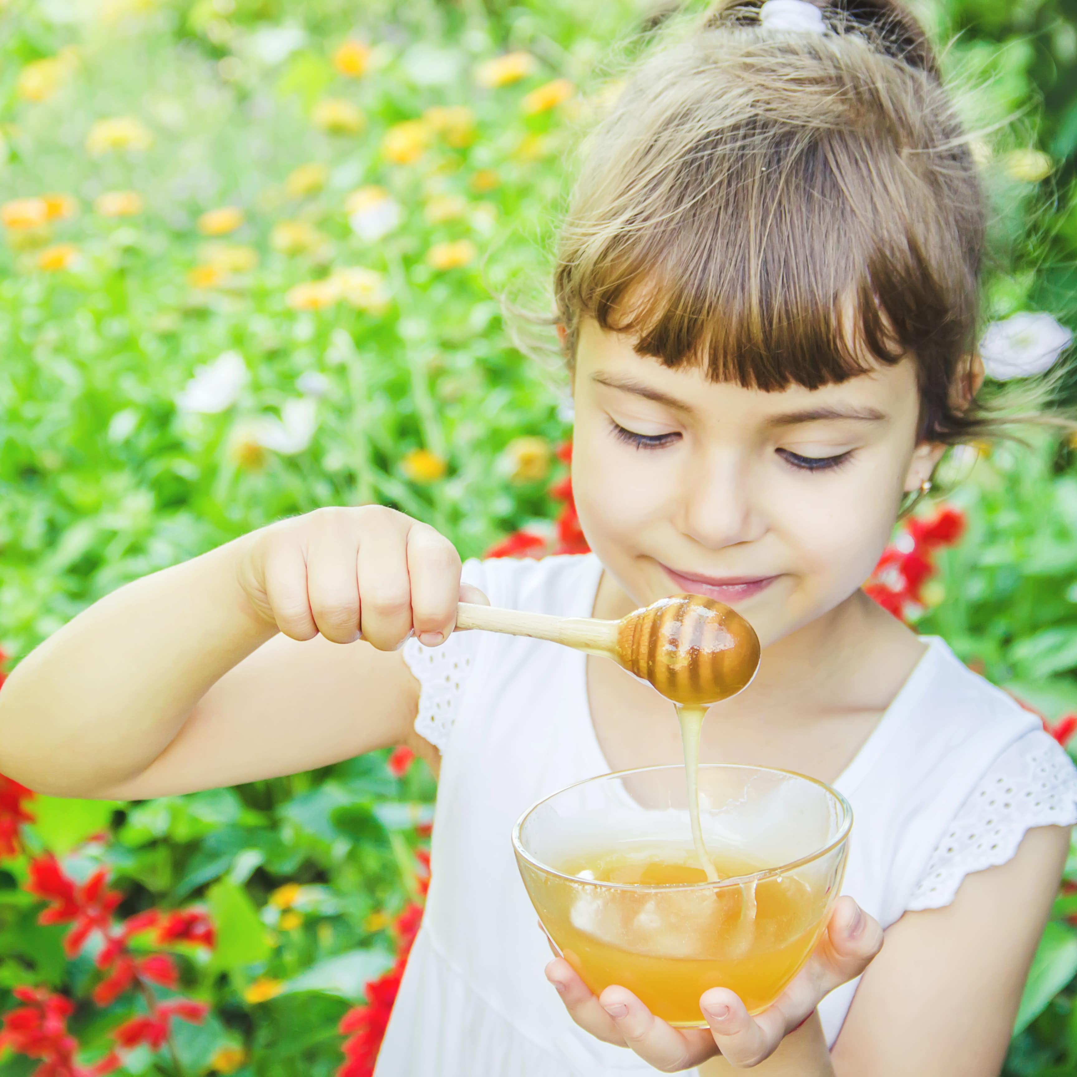 Ein Mädchen mit Zopf hält ein Schälchen Honig und einen Honiglöffel in den Händen. Im Hintergrund Blumen.