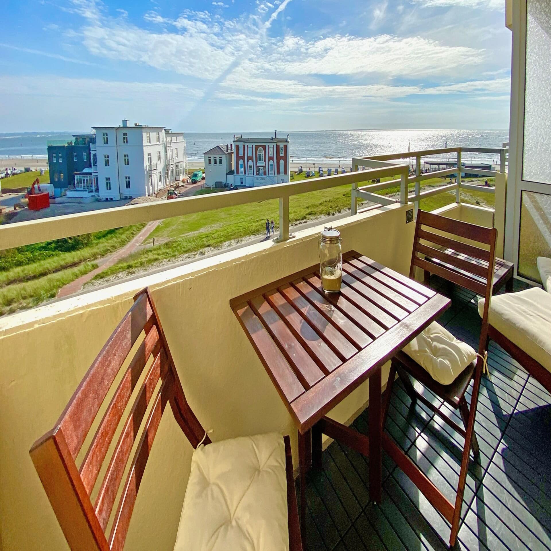 Balkon mit Sitzgelegenheiten und Blick auf Gebäude, den Strand und das Meer. Die Sonne scheint. 