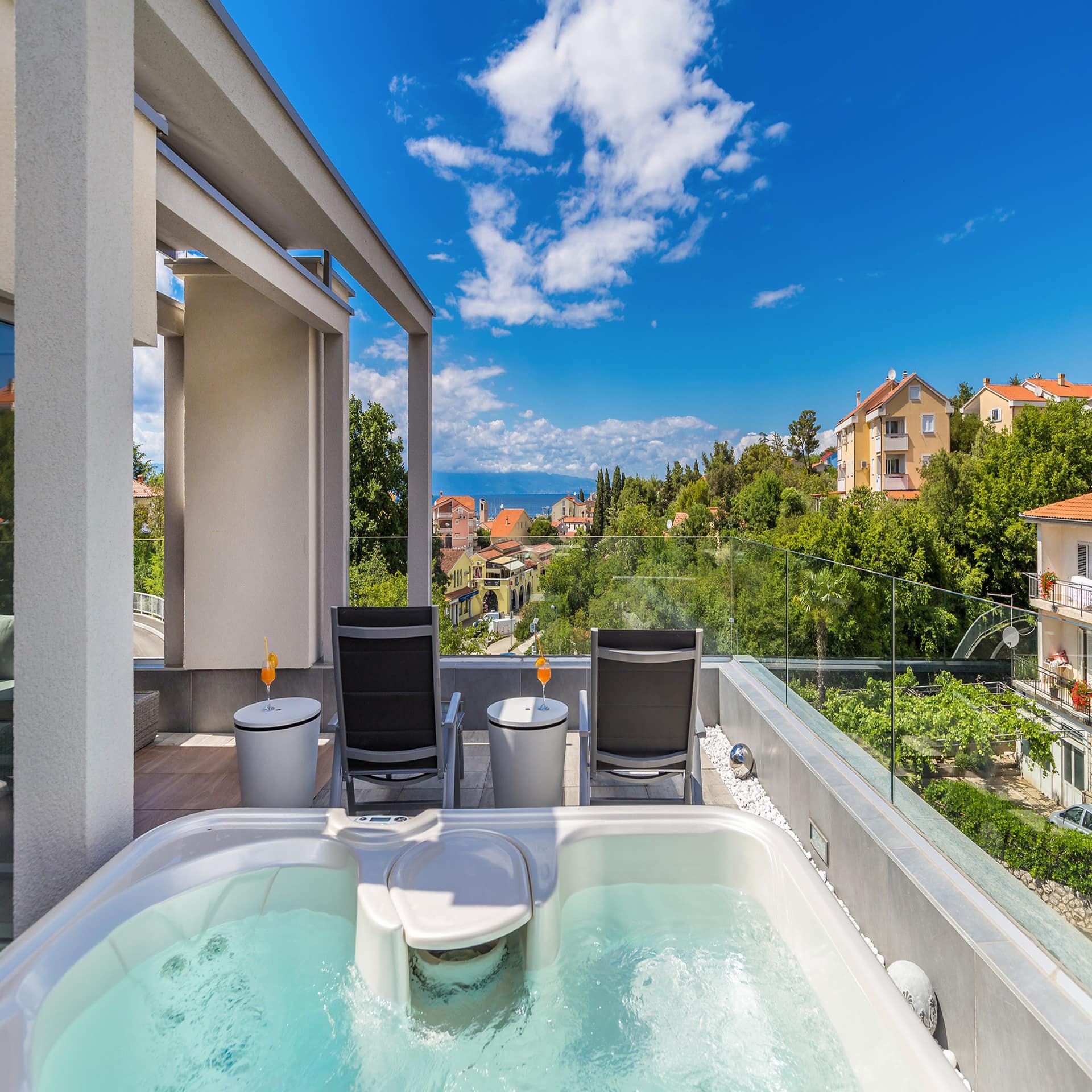 Blick vom Balkon mit Whirlpool und Liegestühlen auf den Ort, Bäume und das Meer. Blauer Himmel und Sonnenschein. 