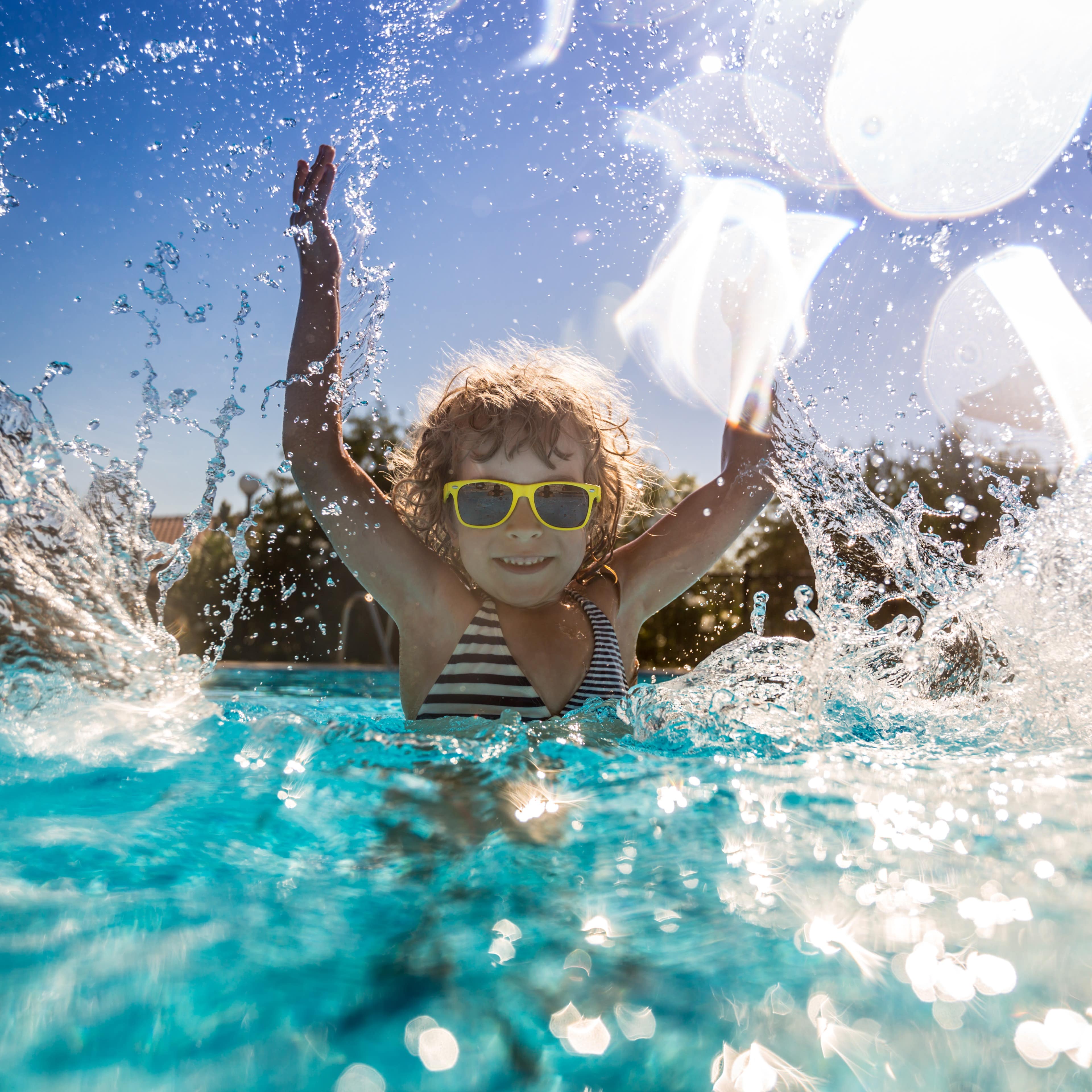Mädchen mit gestreiftem Badeanzug und gelber Sonnenbrille spritzt Wasser in einem Schwimmbecken.