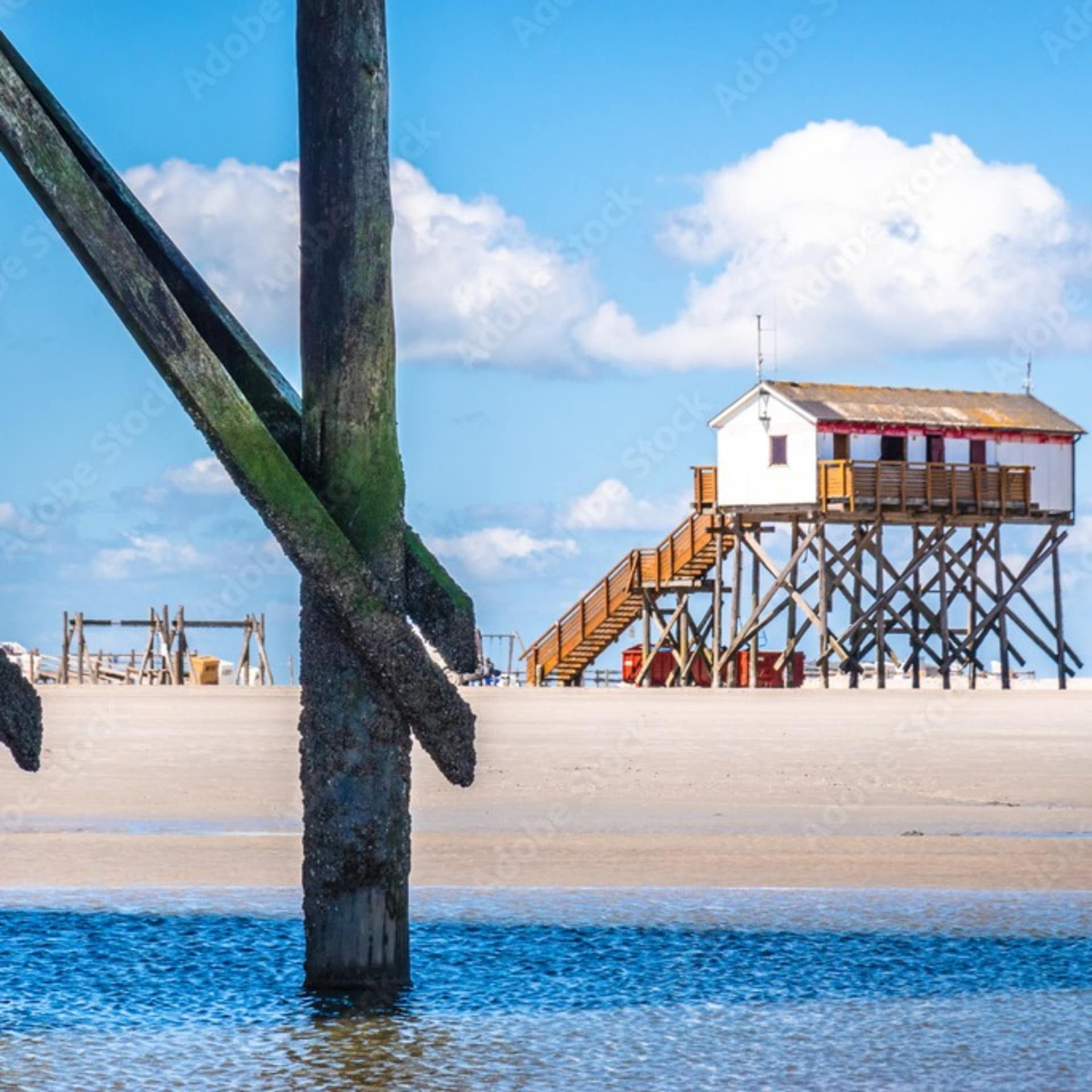 Blick an einigen Pfählen vorbei auf den Strand und ein Haus auf Pfählen.