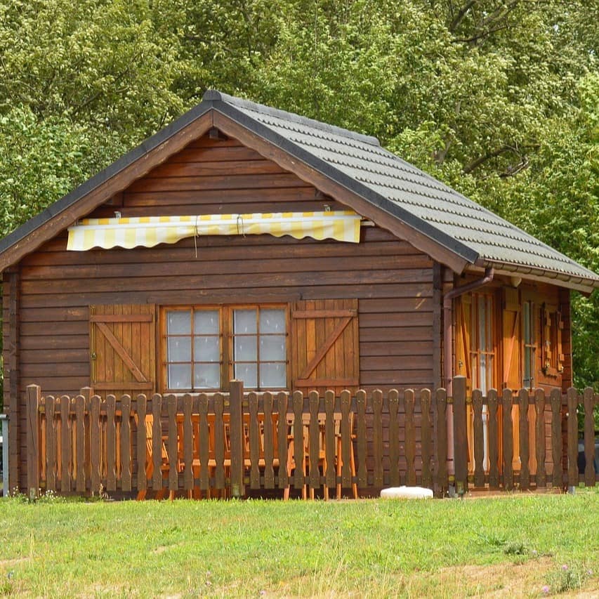 Eine kleine Waldhütte mit gelb-weiß gestreifter Markise und Gartenzaun.
