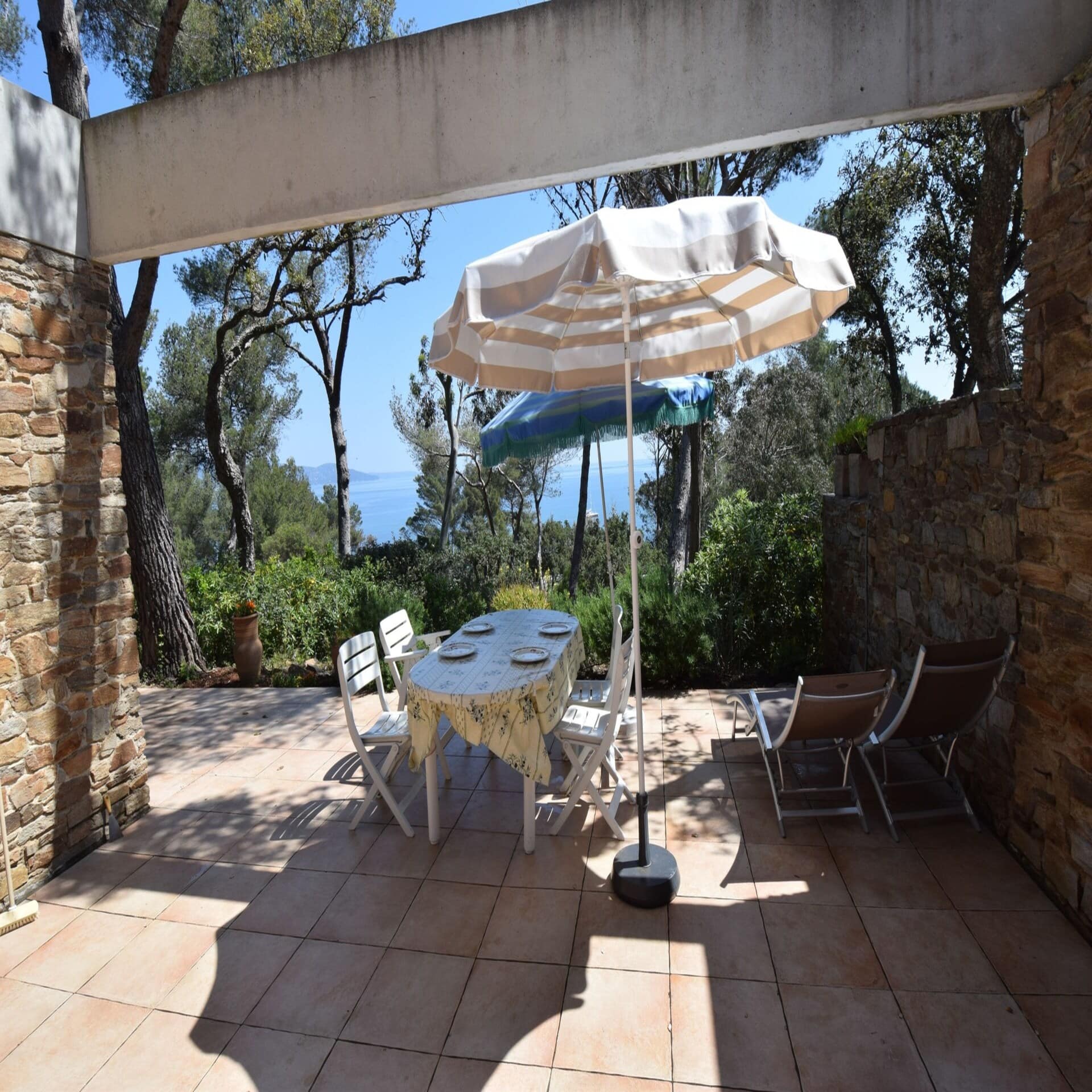 Terrasse mit Sitzgelegenheiten, Liegen und Sonnenschirmen sowie Blick durch Bäume auf das Wasser.