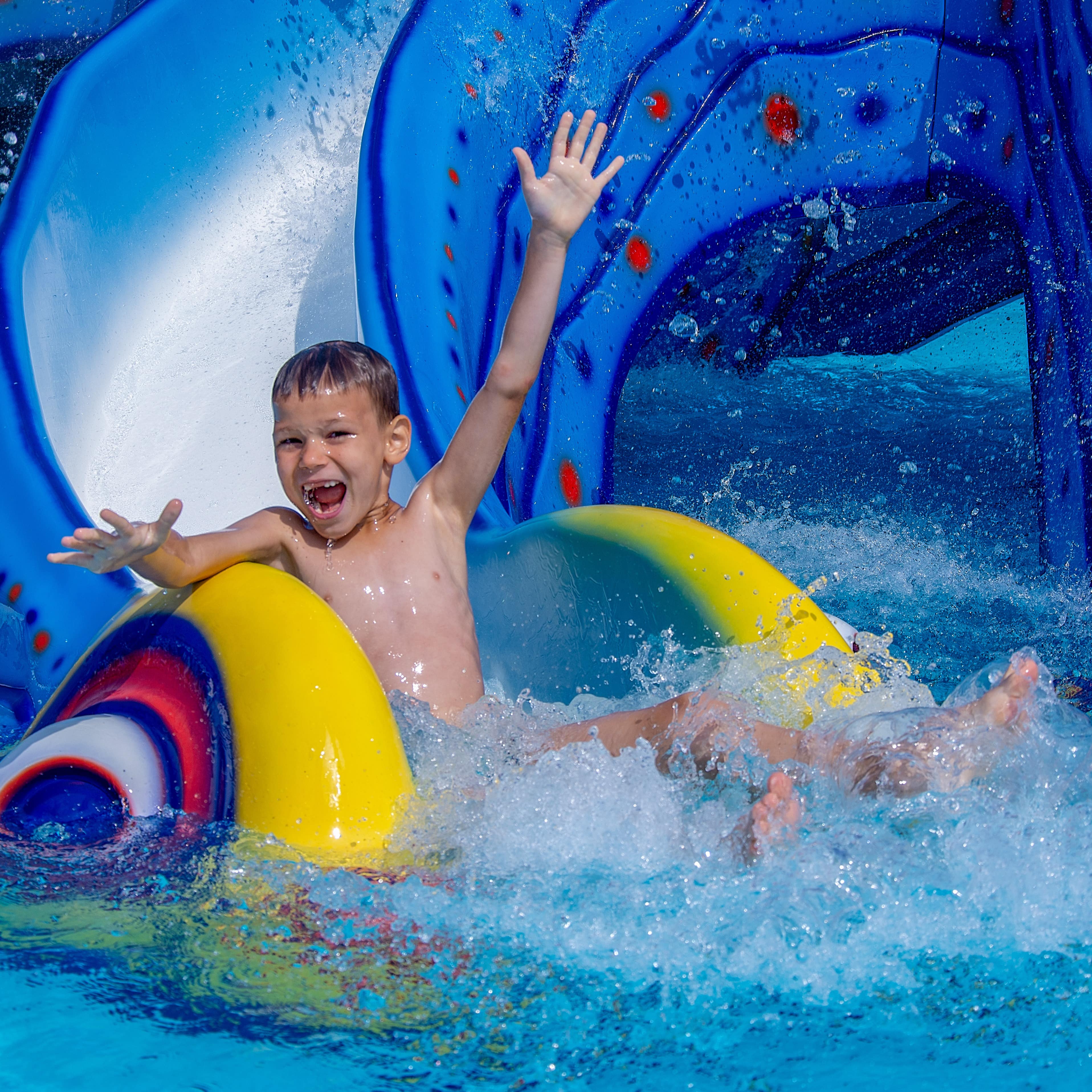 Junge mit hochgestreckten Armen auf einer blauen Wasserrutsche im Wasserpark.