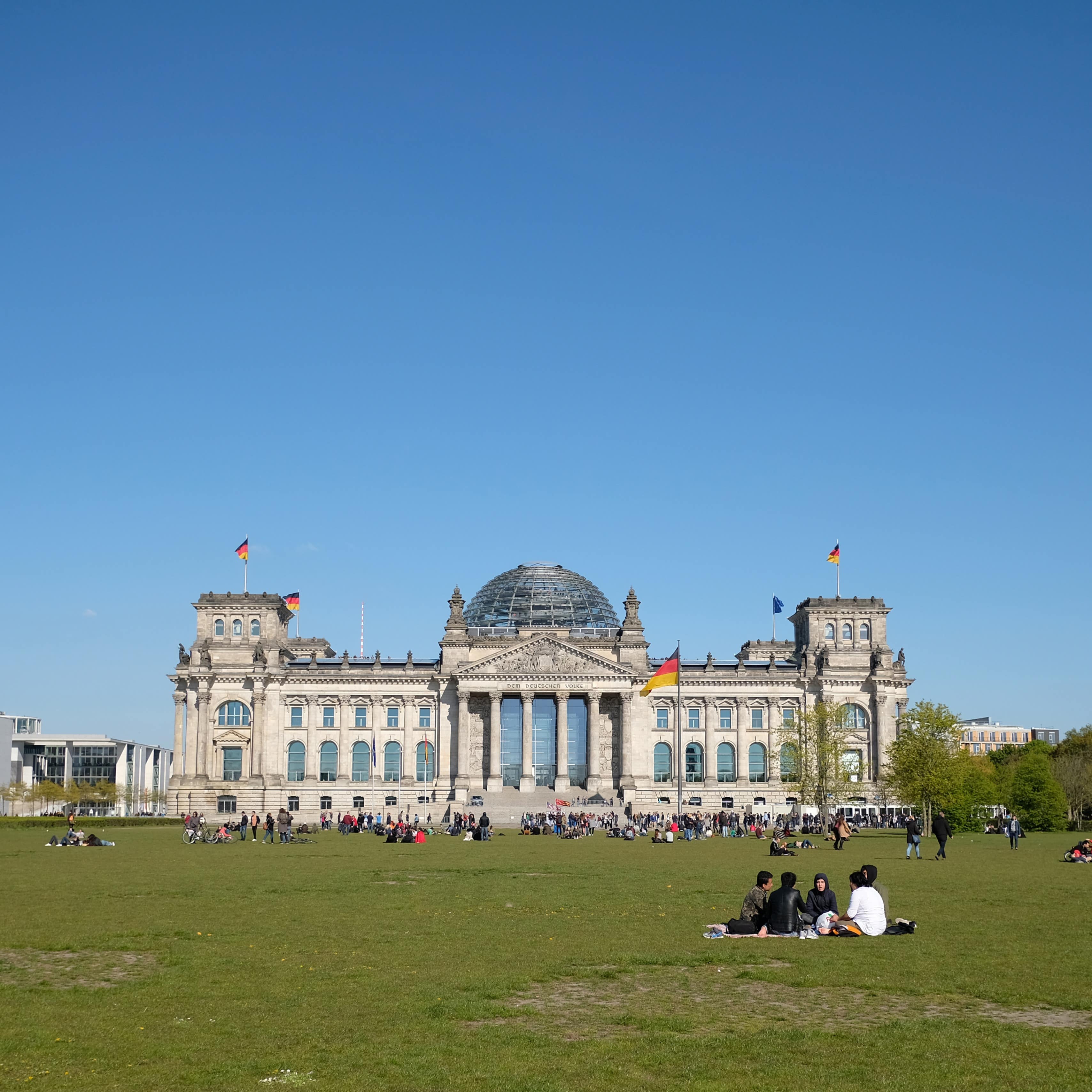 Panoramablick: Verschiedene kleine Gruppen sitzen auf der Wieder vor dem Reichstag und picknicken in der Sonne.