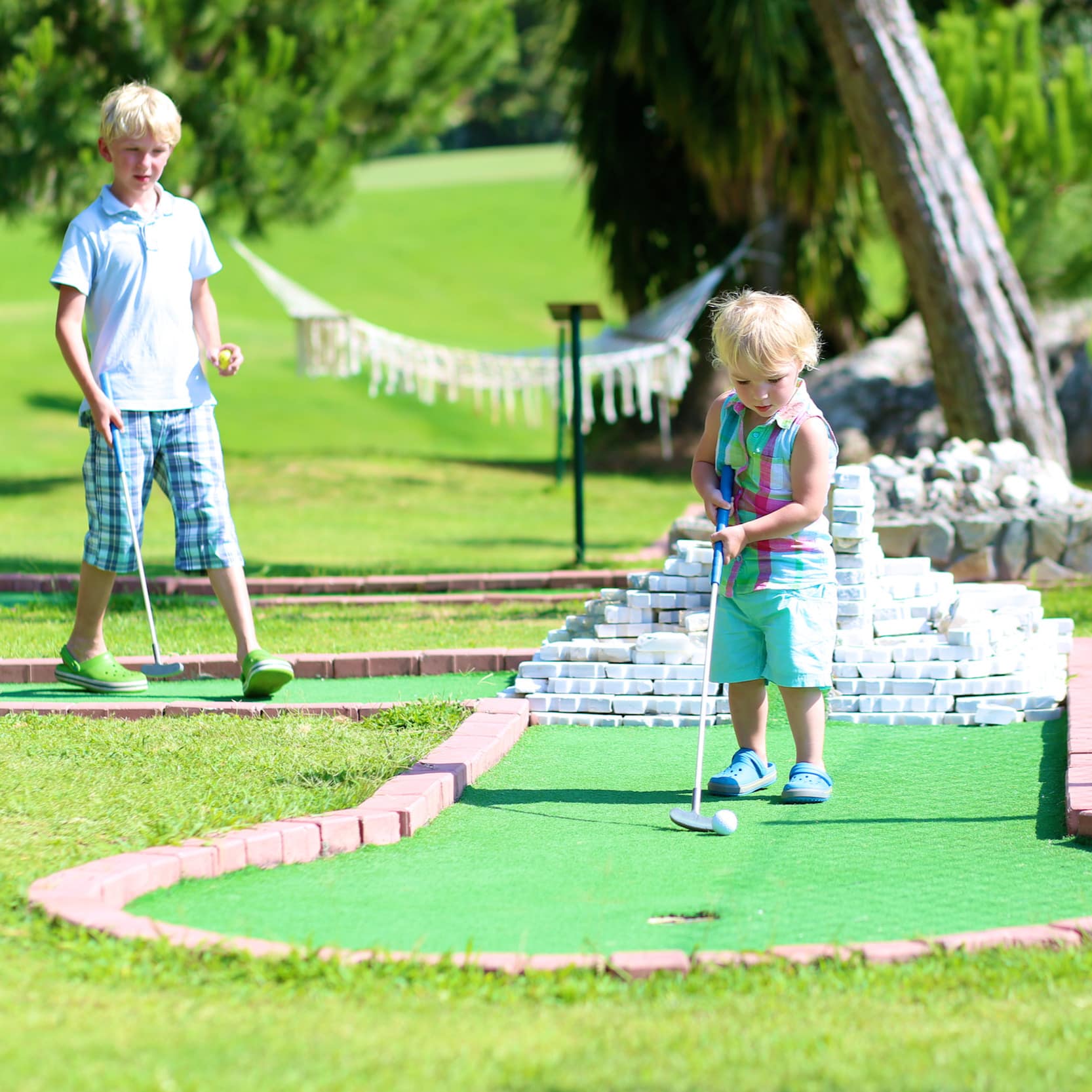 Junge und 1 Kleinkind in Sommerkleidung beim Minigolf spielen.