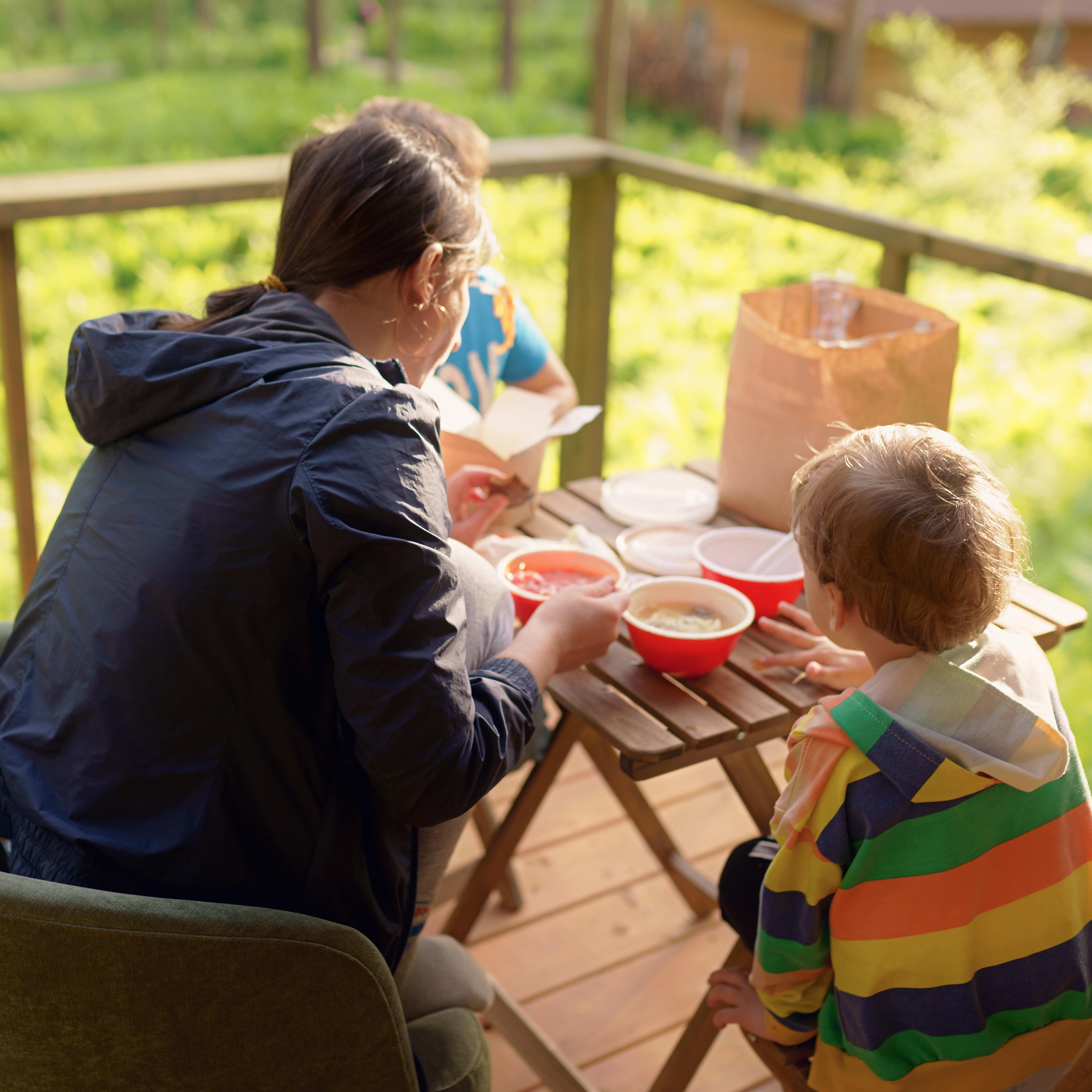 Mutter und 2 Kinder sitzen auf einer Holzterrasse einer Glamping-Unterkunft und Essen.