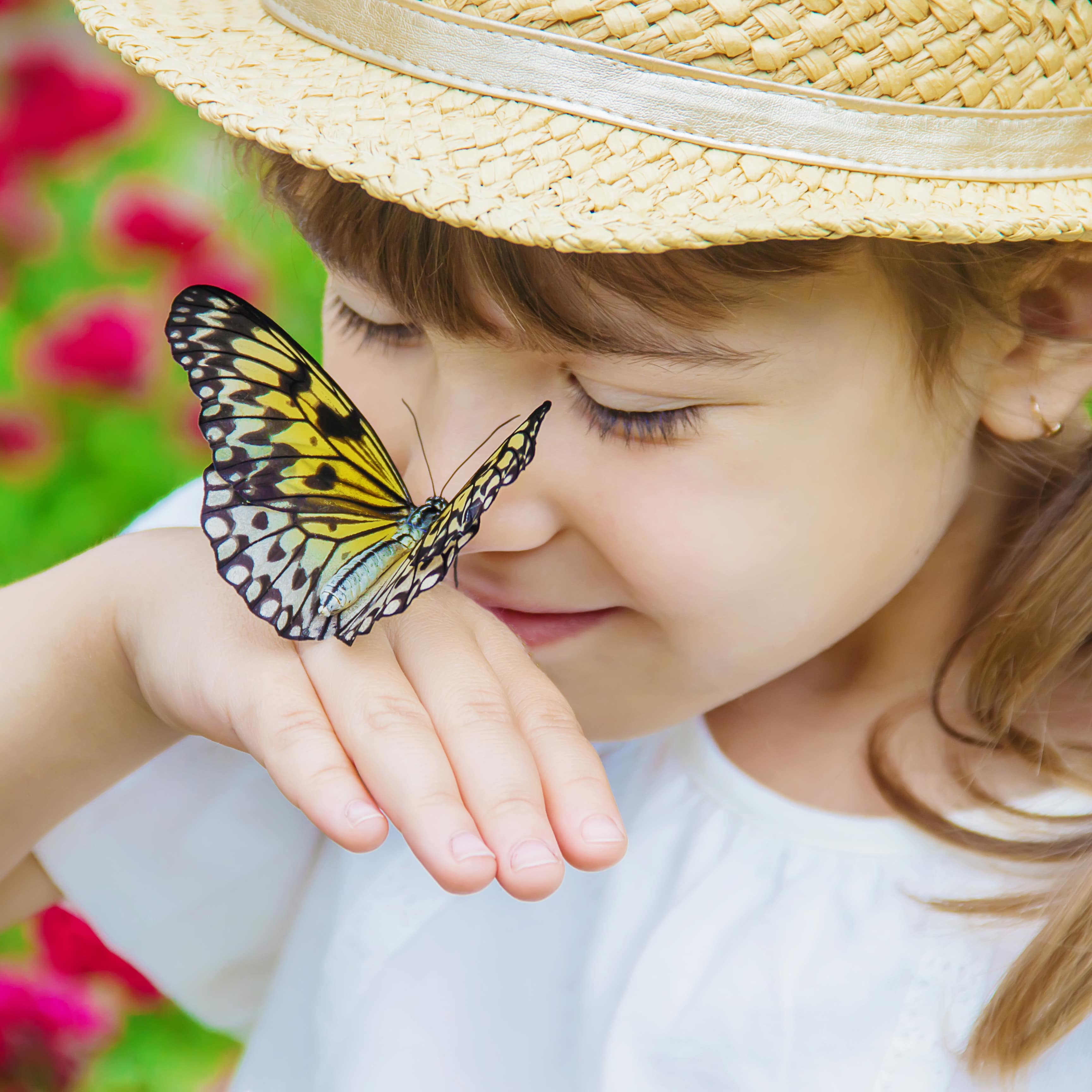 Mädchen in weißem T-Shirt und mit Strohhut hat einen Schmetterling auf der Hand und hält ihm die Nase hin.