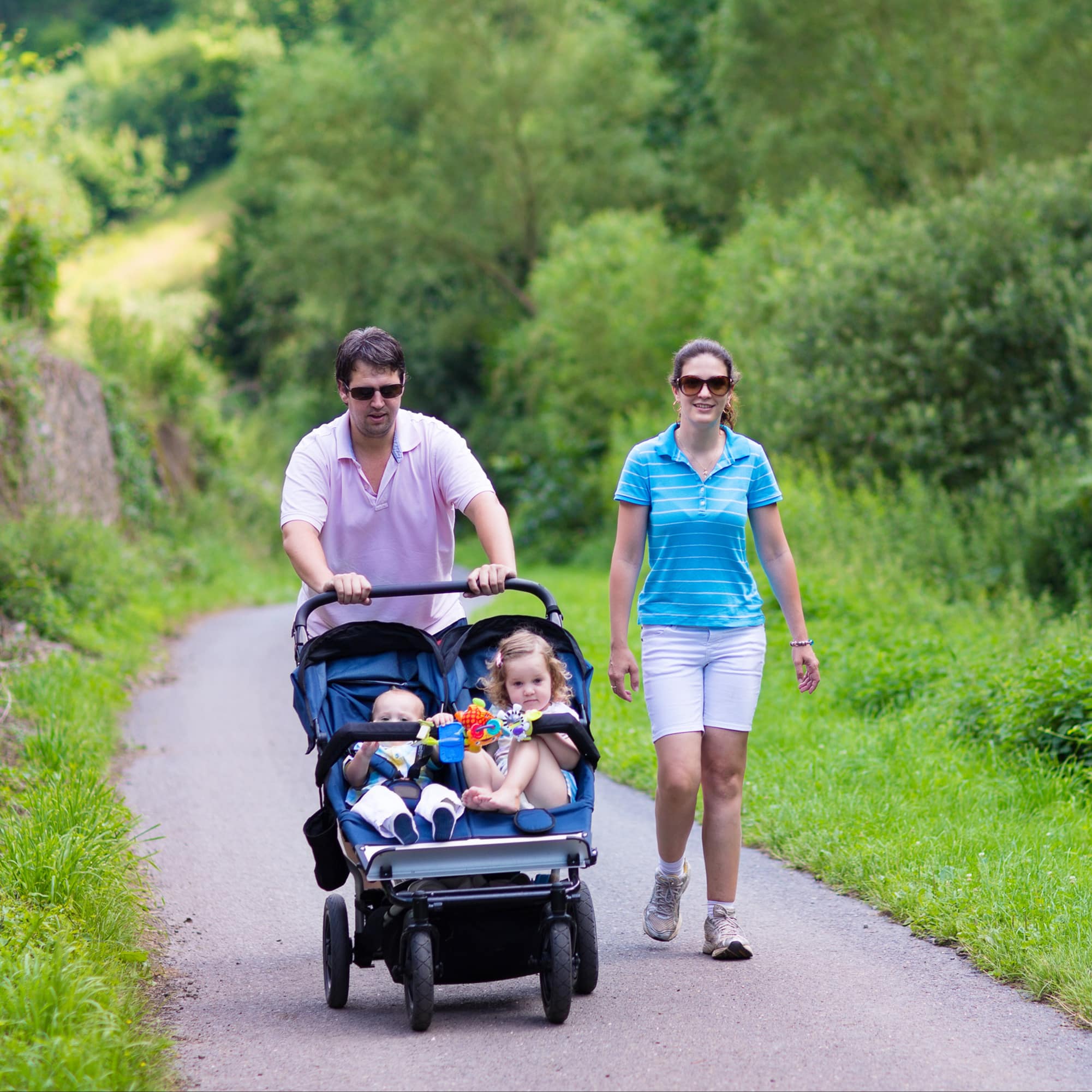 Familie beim Wandern: Der Vater schiebt einen Zwillingskinderwagen mit Baby und Kleinkind.