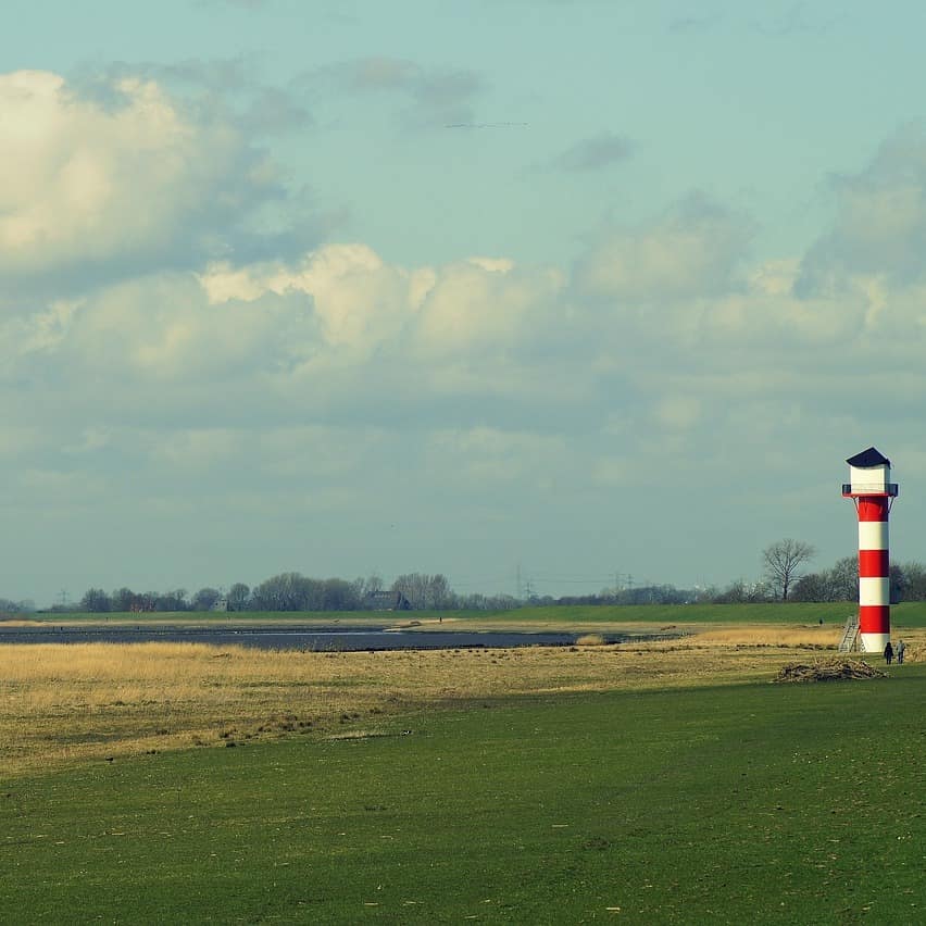 Ein Leuchtturm steht auf einer Wiese am Wasser, im Hintergrund sind ein Haus und Windräder zu sehen.