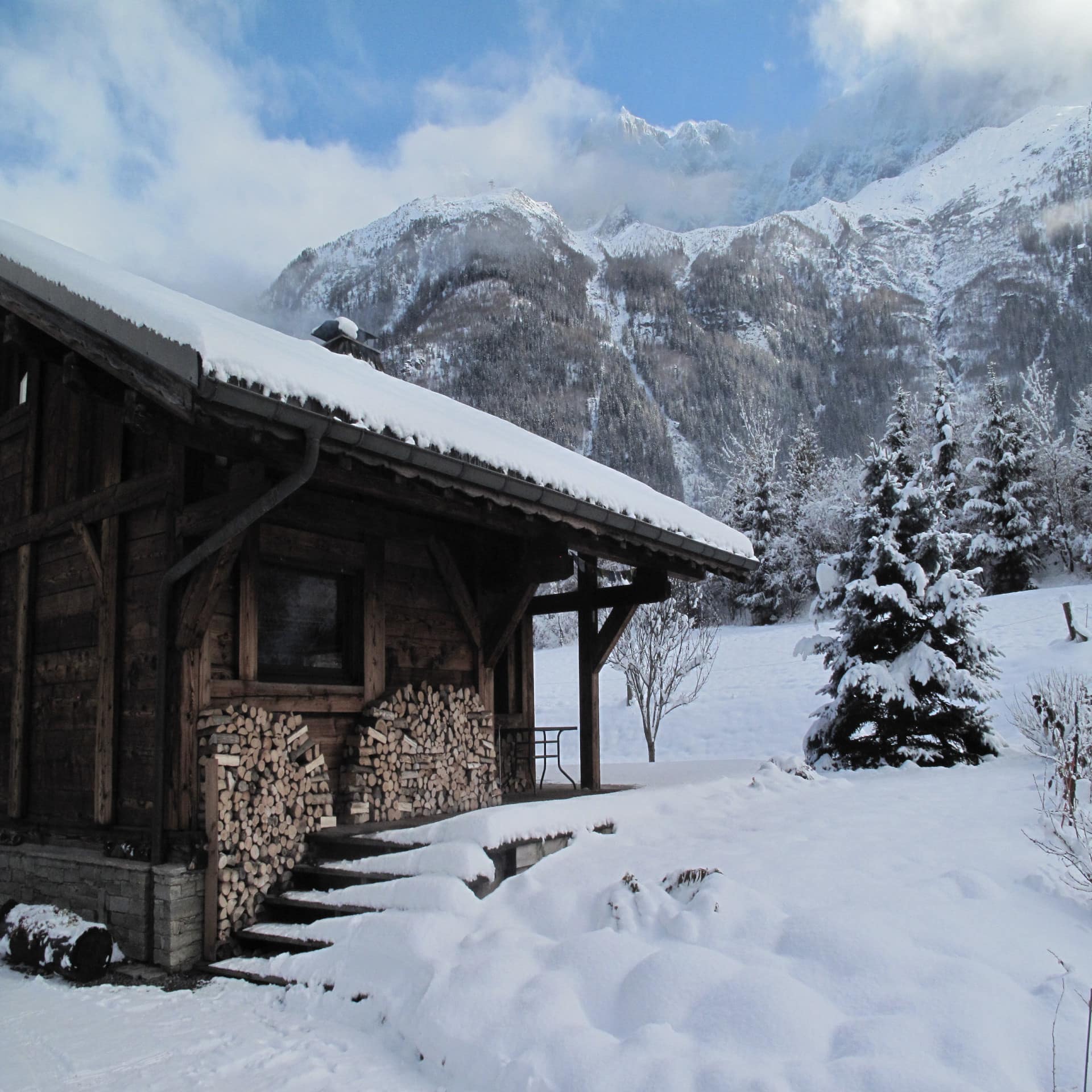 Verschneite Holzhütte mit Feuerholz daneben, dahinter die Berge von Chamonix.