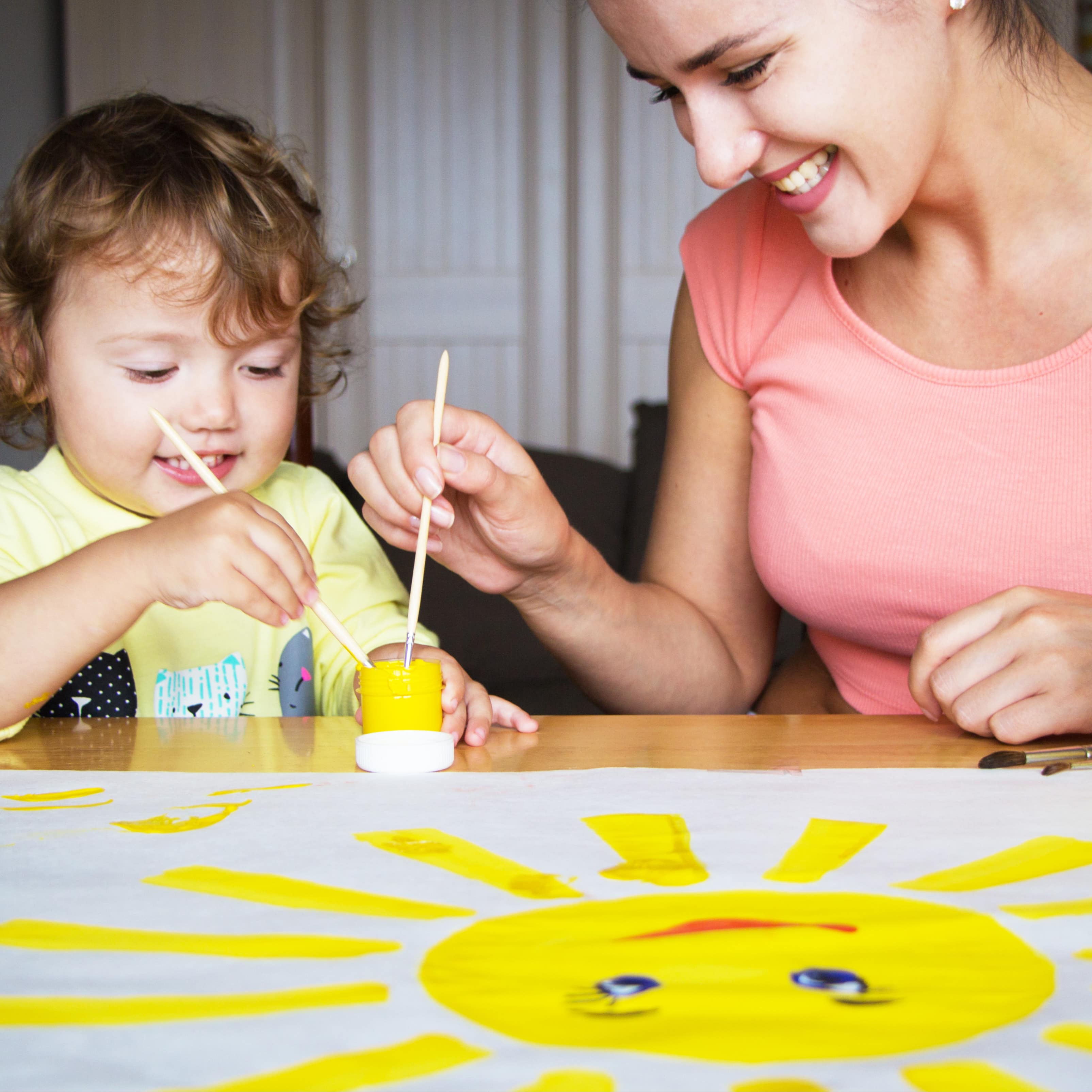 Babysitterin malt mit einem Kleinkind eine gelbe Sonne auf einem großen Blatt.