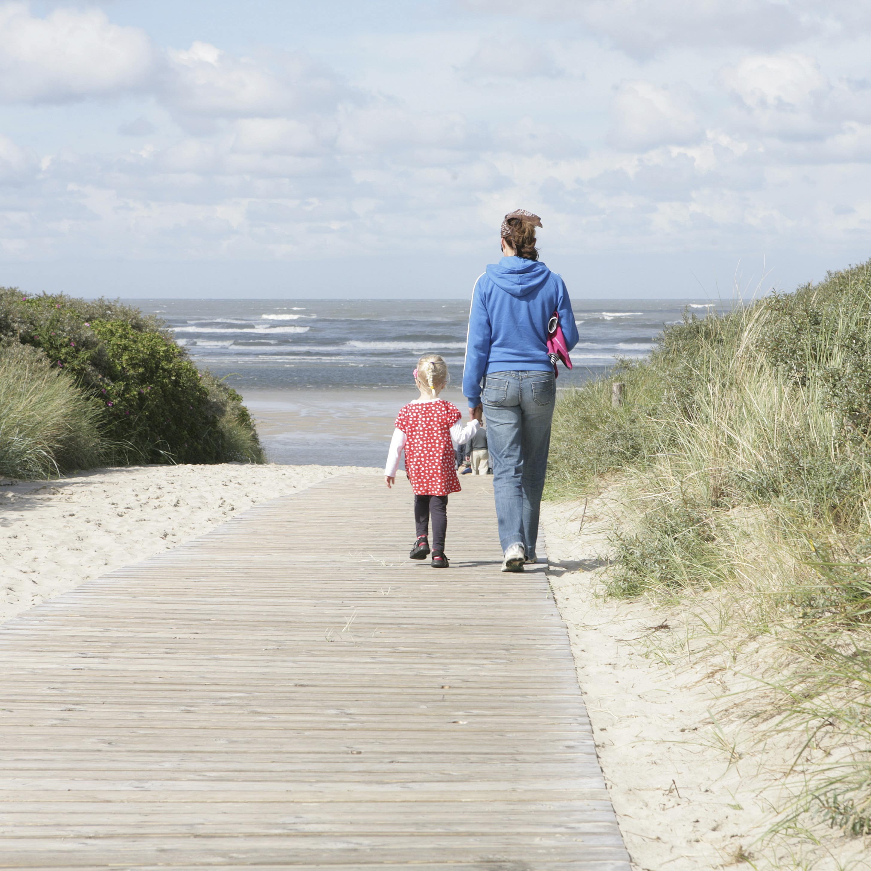 Rückenansicht: Mutter und Tochter laufen einen Holzsteg entlang auf den Strand zu.