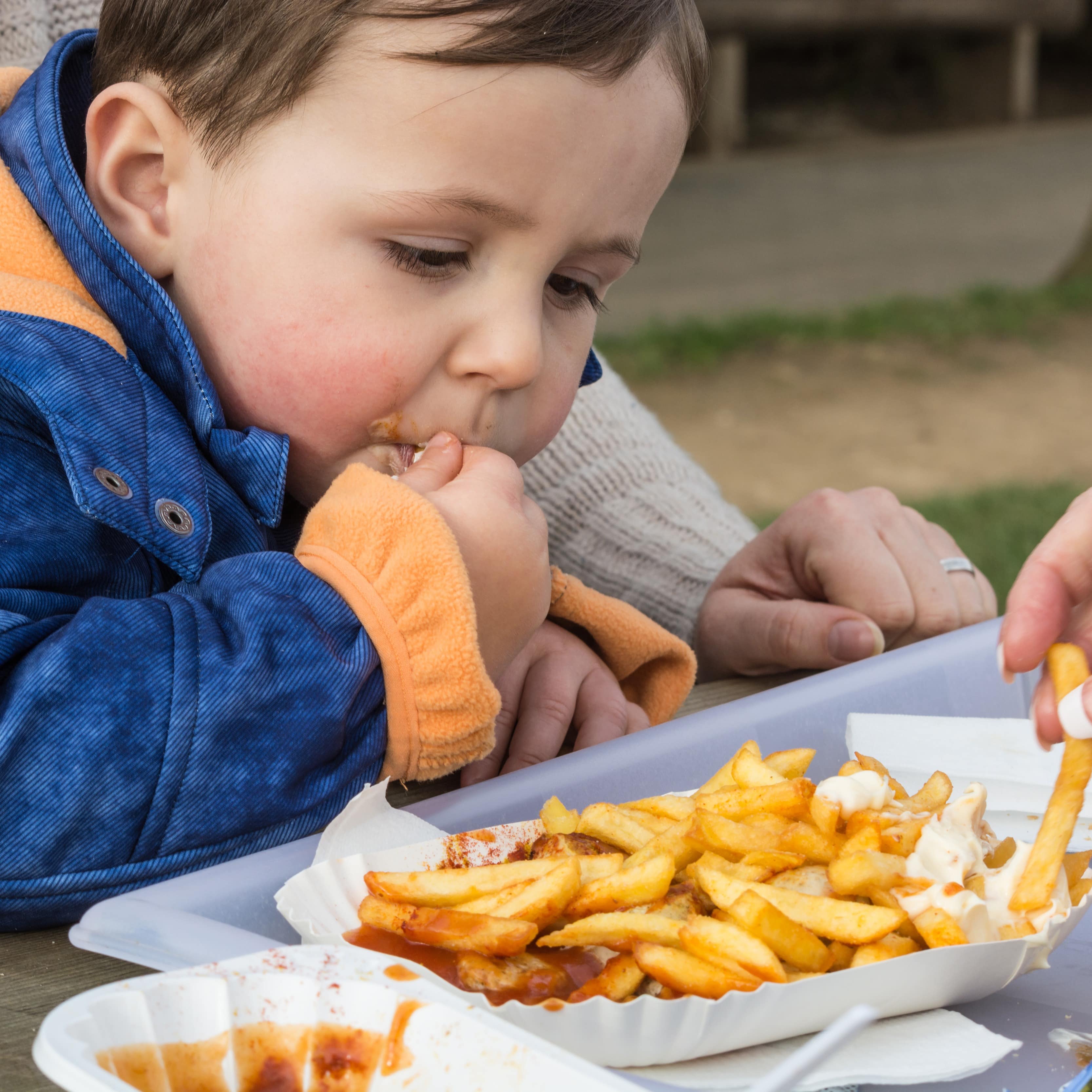 Ein kleiner June auf dem Schoß seiner Mutter isst Pommes und Currywurst mit Ketchup und Majo.