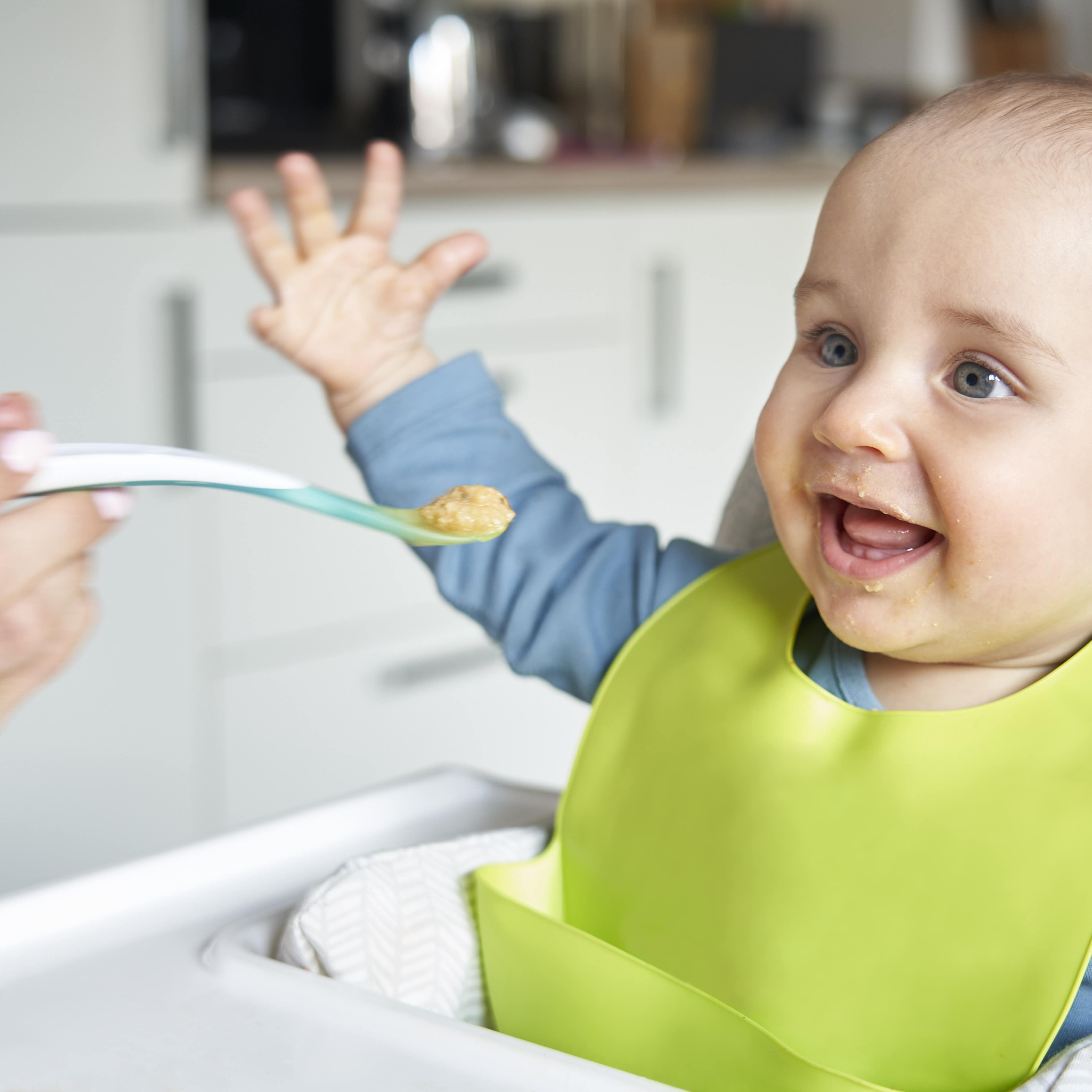 Baby in blauem Top mit grünem Lätzchen freut sich, dass es von der Mutter gefüttert wird.