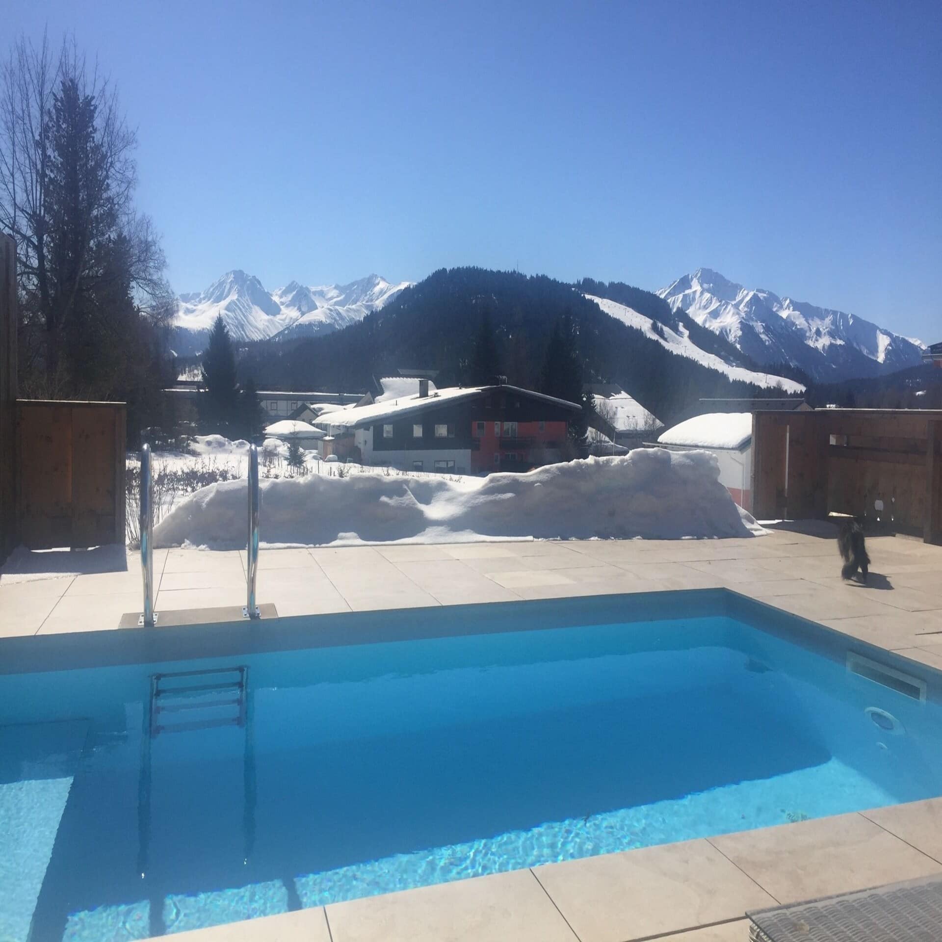 Verschneiter Garten mit Pool und Blick auf die Berge. Die Sonne scheint. 