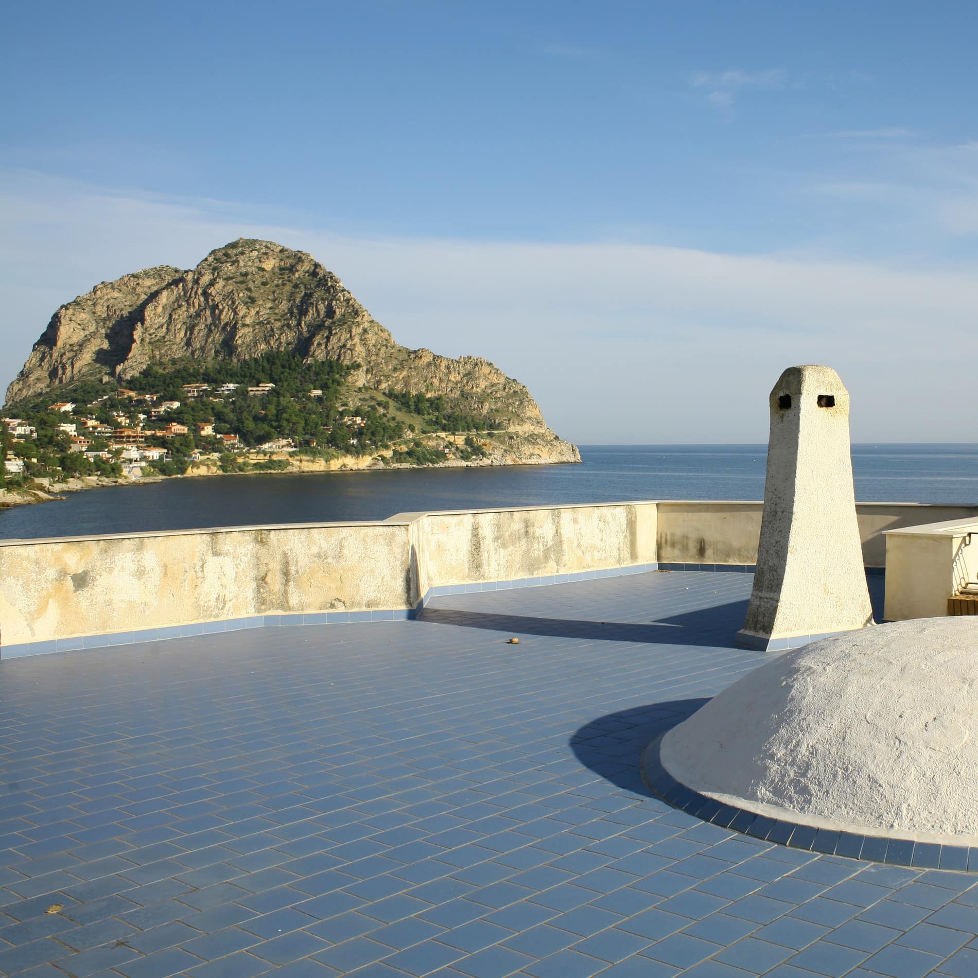 Über der blauen Terrasse der Ferienwohnung in Sizilien erhebt sich der charakteristische Felsen des Capo Zafferano aus dem Meer