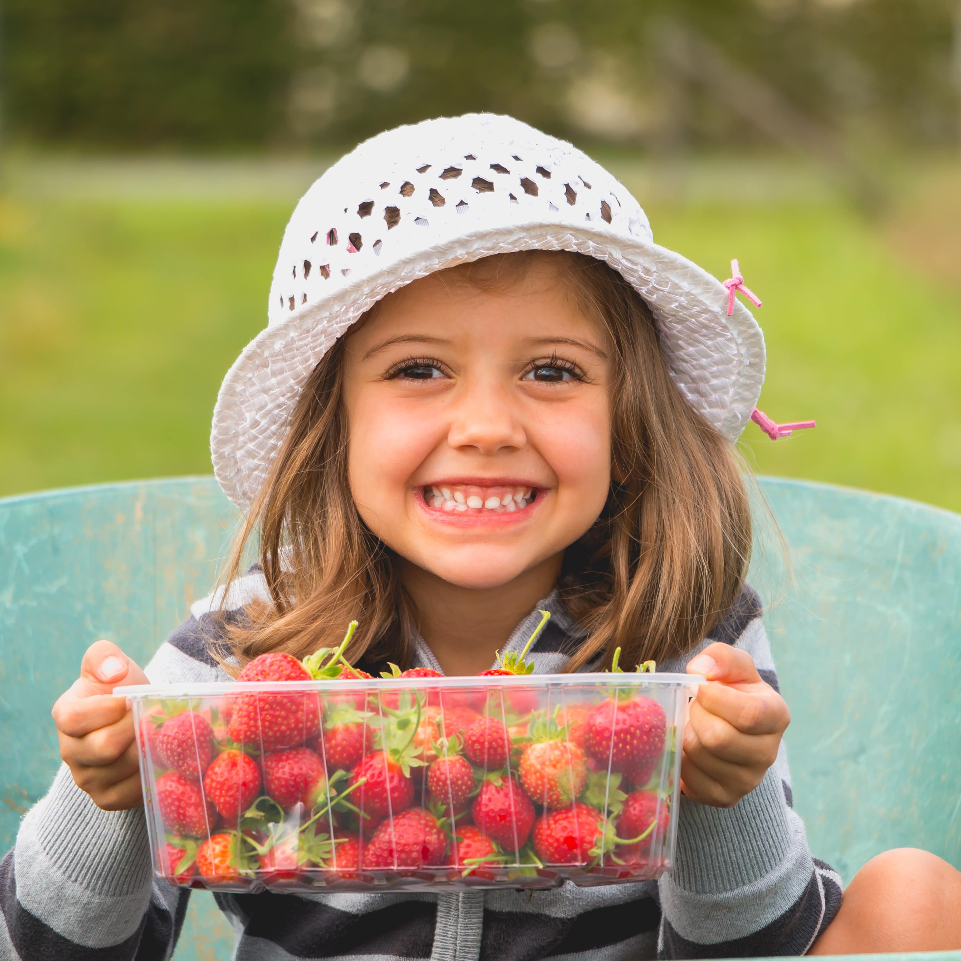 Mädchen mit weißem Strohhut hält eine Plastikschale mit Erdbeeren in der Hand und strahlt in die Kamera.
