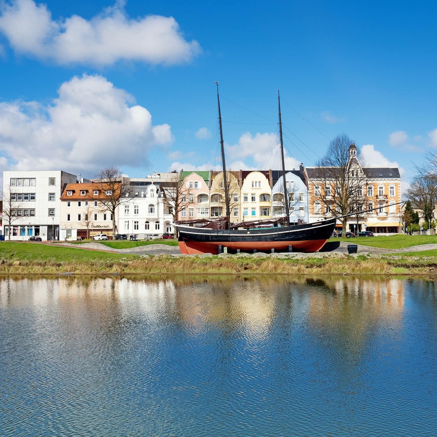 Blick vom Wasser auf ein ausgestelltes Segelboot an Land mit Häusern im Hintergrund
