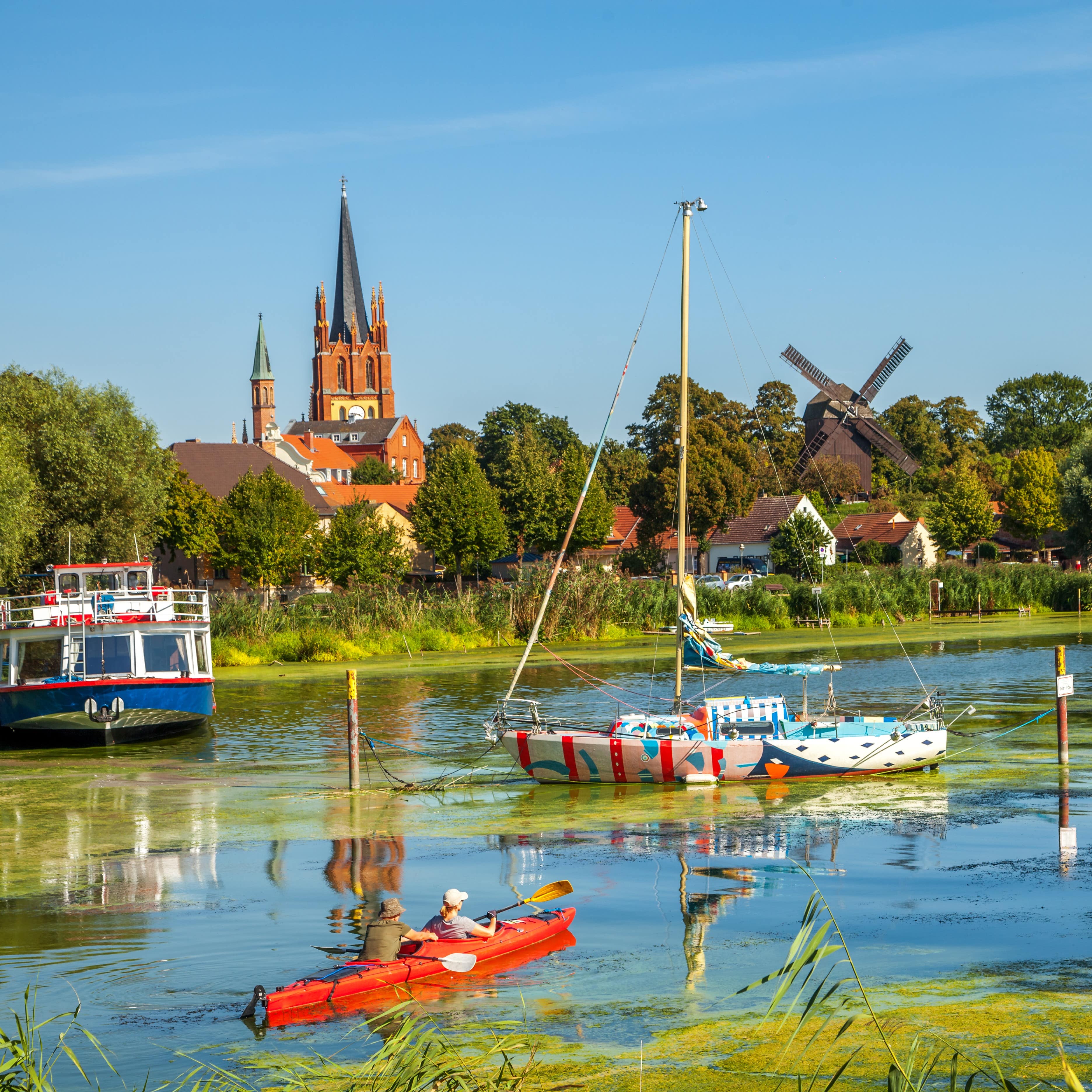 Blick auf die Havel mit Kanufahrern und Booten, am Ufer die Stadt Werder, eine Kirche, eine Windmühle und Bäume.