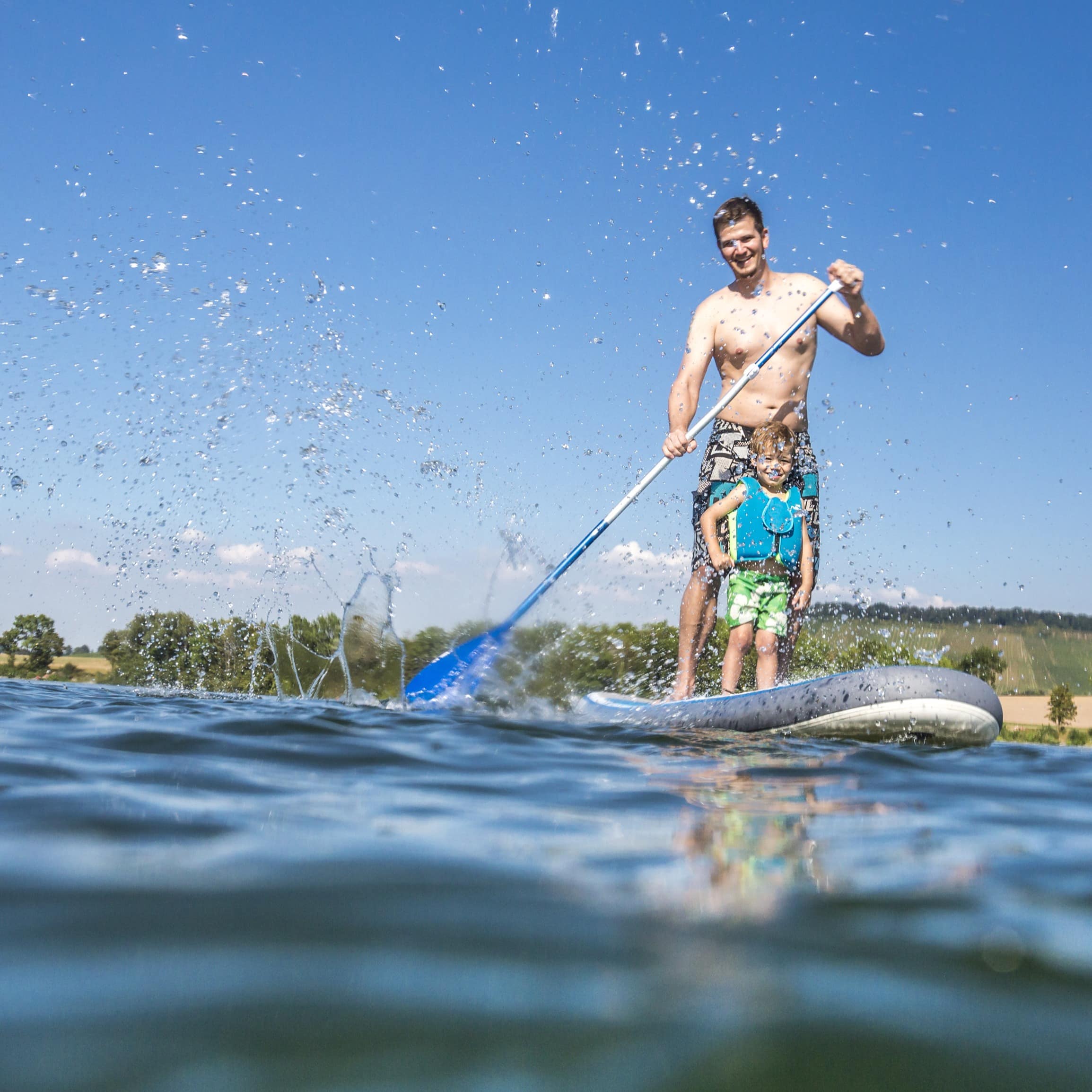 Vater und Sohn auf einem SUP auf dem See. Der Junge trägt eine Schwimmweste.