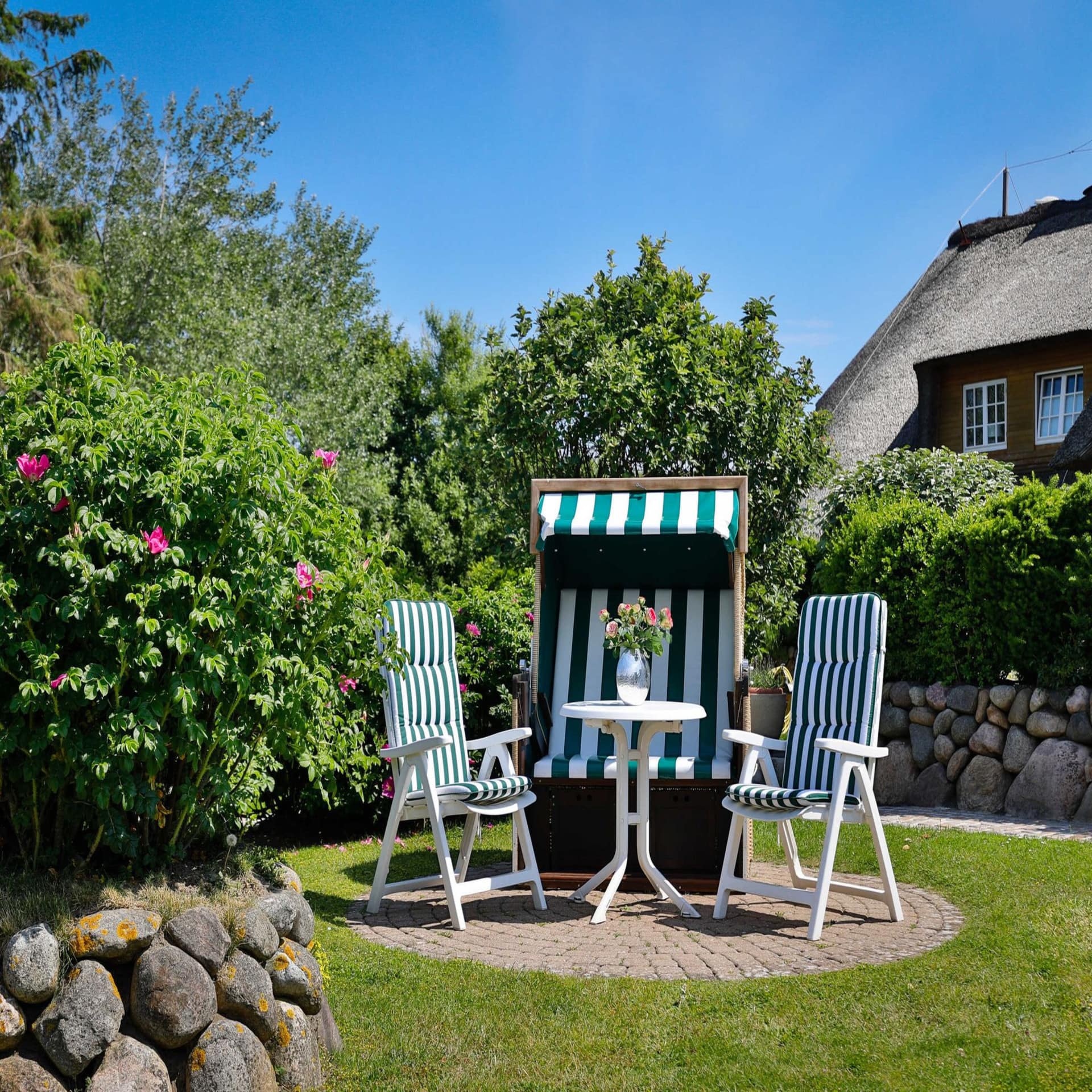 Ein grün-weiß gestreifter Strandkorb, 2 Stühle und 1 Tisch mit Blumen im Garten bei Sonnenschein.