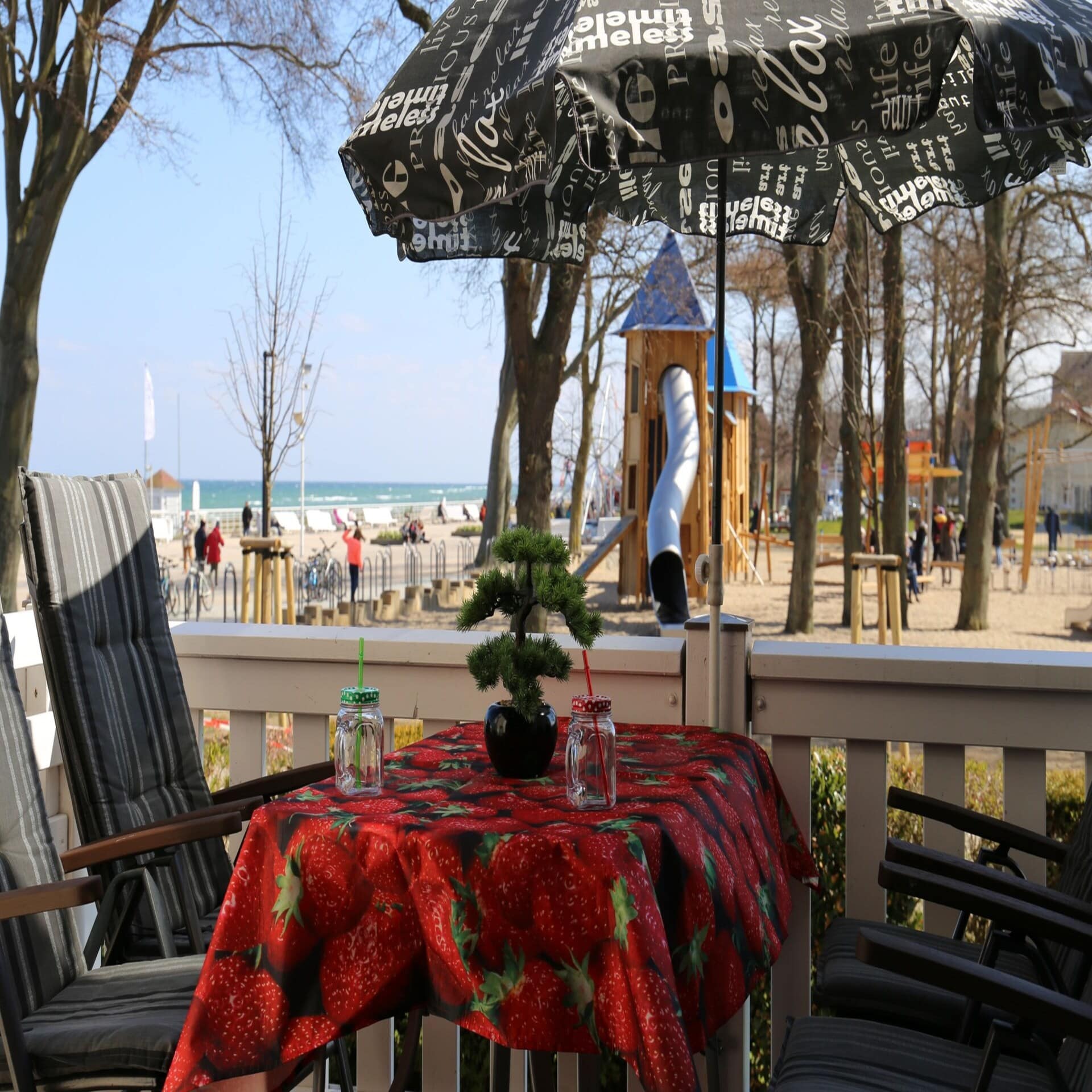 Veranda mit Tisch mit Erdbeer-Tischdecke und 4 Stühlen und Blick auf einen Spielplatz an der Ostseepromenade.