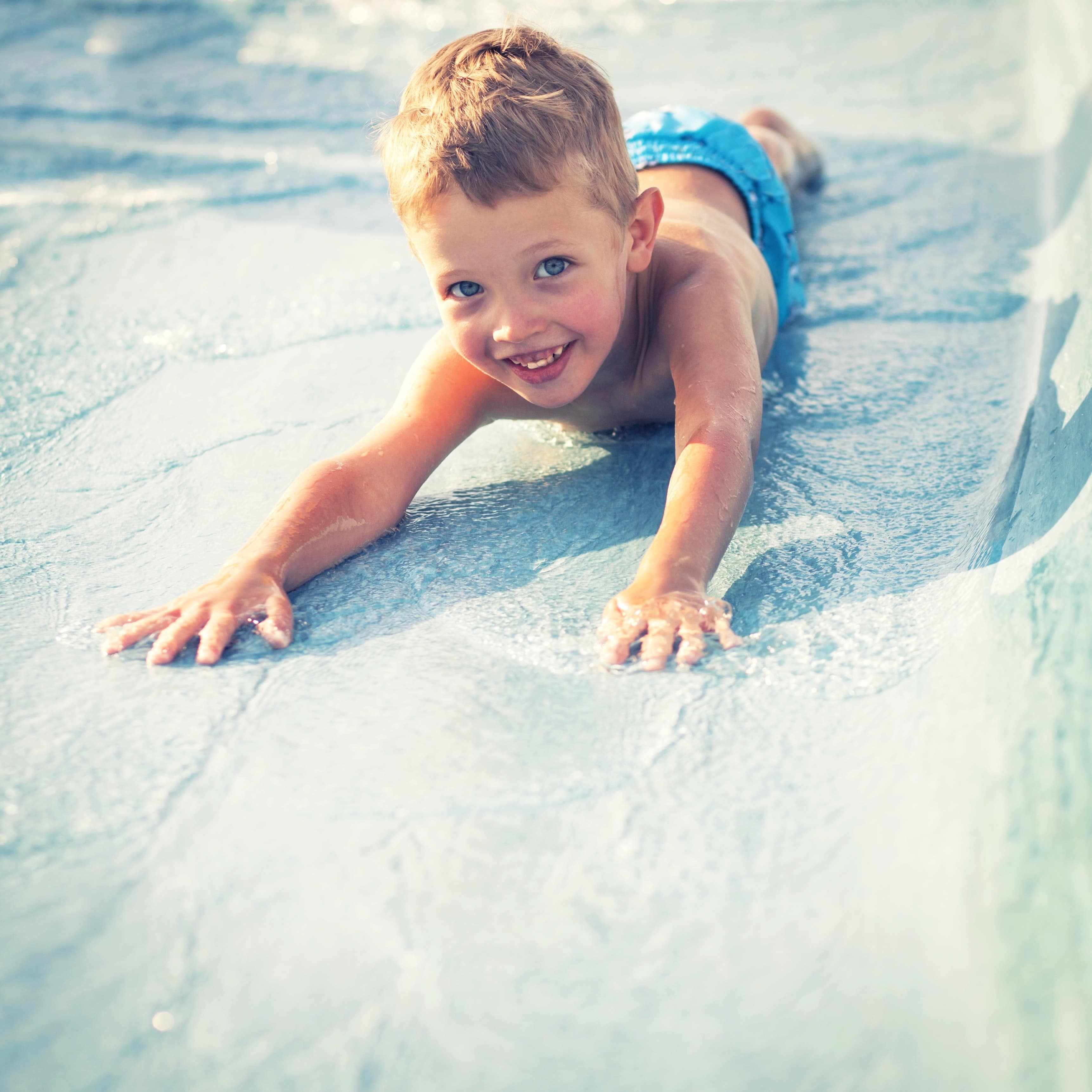 Blonder Junge mit blauen Augen rutscht bäuchlings eine Wasserrutsche hinunter und lacht in die Kamera.