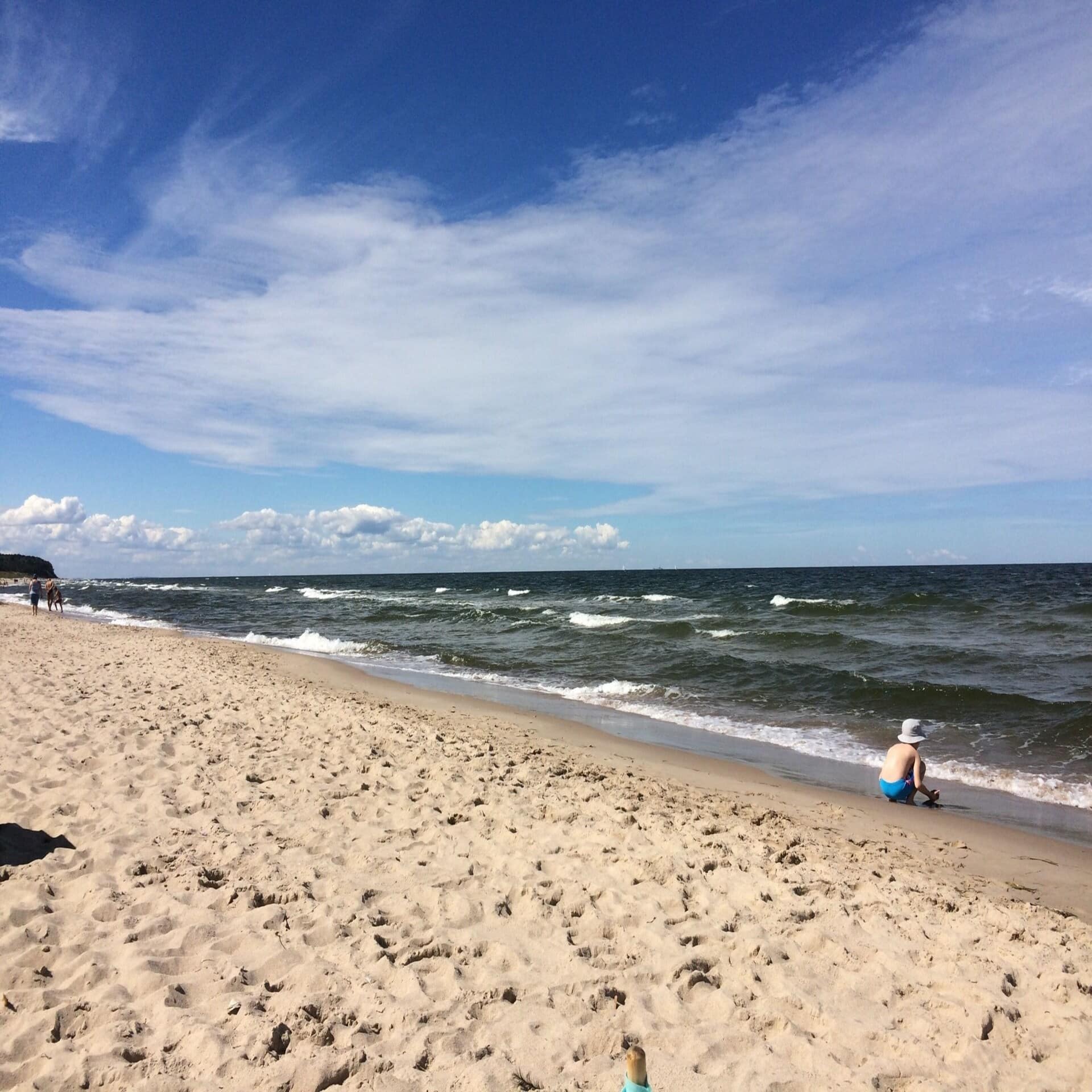 Ein kleiner Junge mit blauer Badehose und weißem Hut spielt am Strand am Meer.