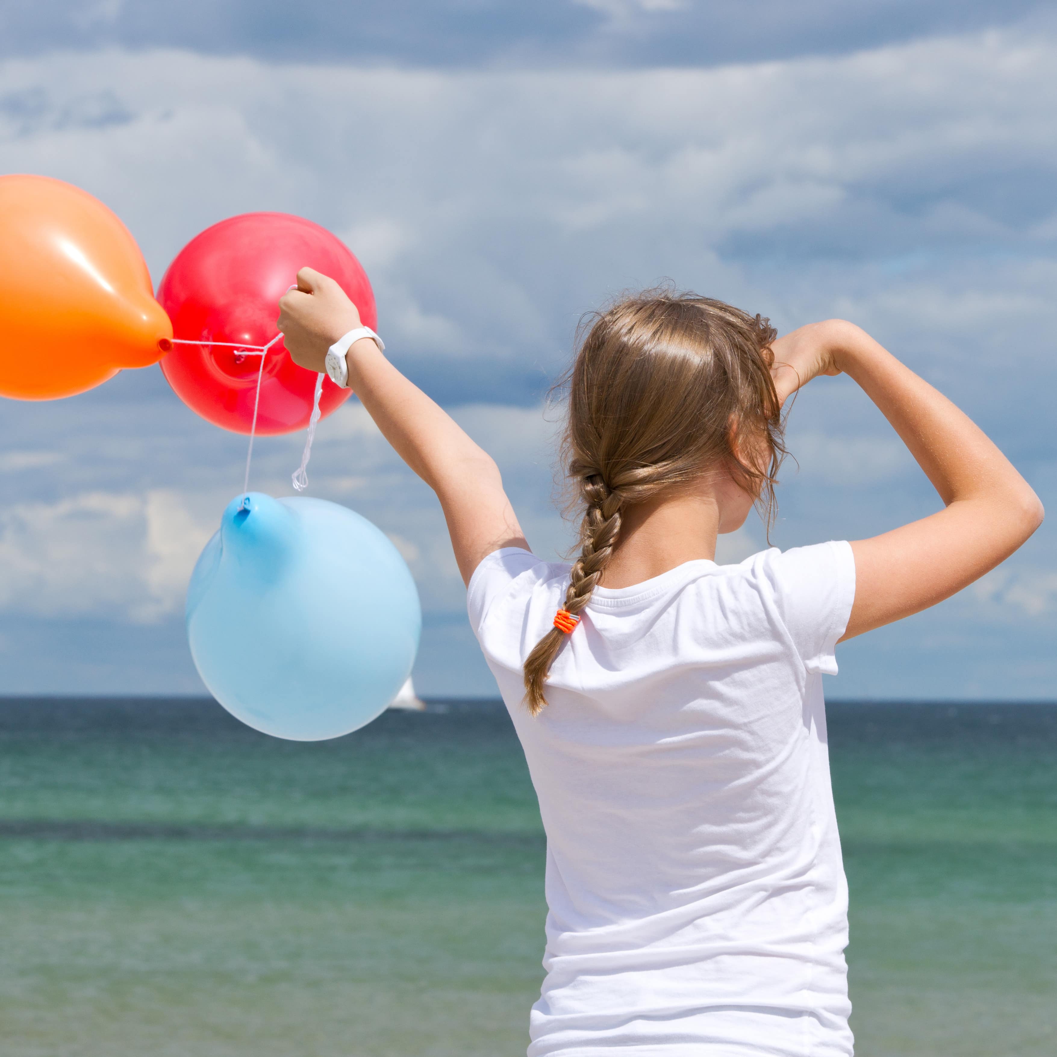 Rückenansicht: Mädchen in weißem T-Shirt mit 2 Ballons in der Hand schaut aufs Meer hinaus.