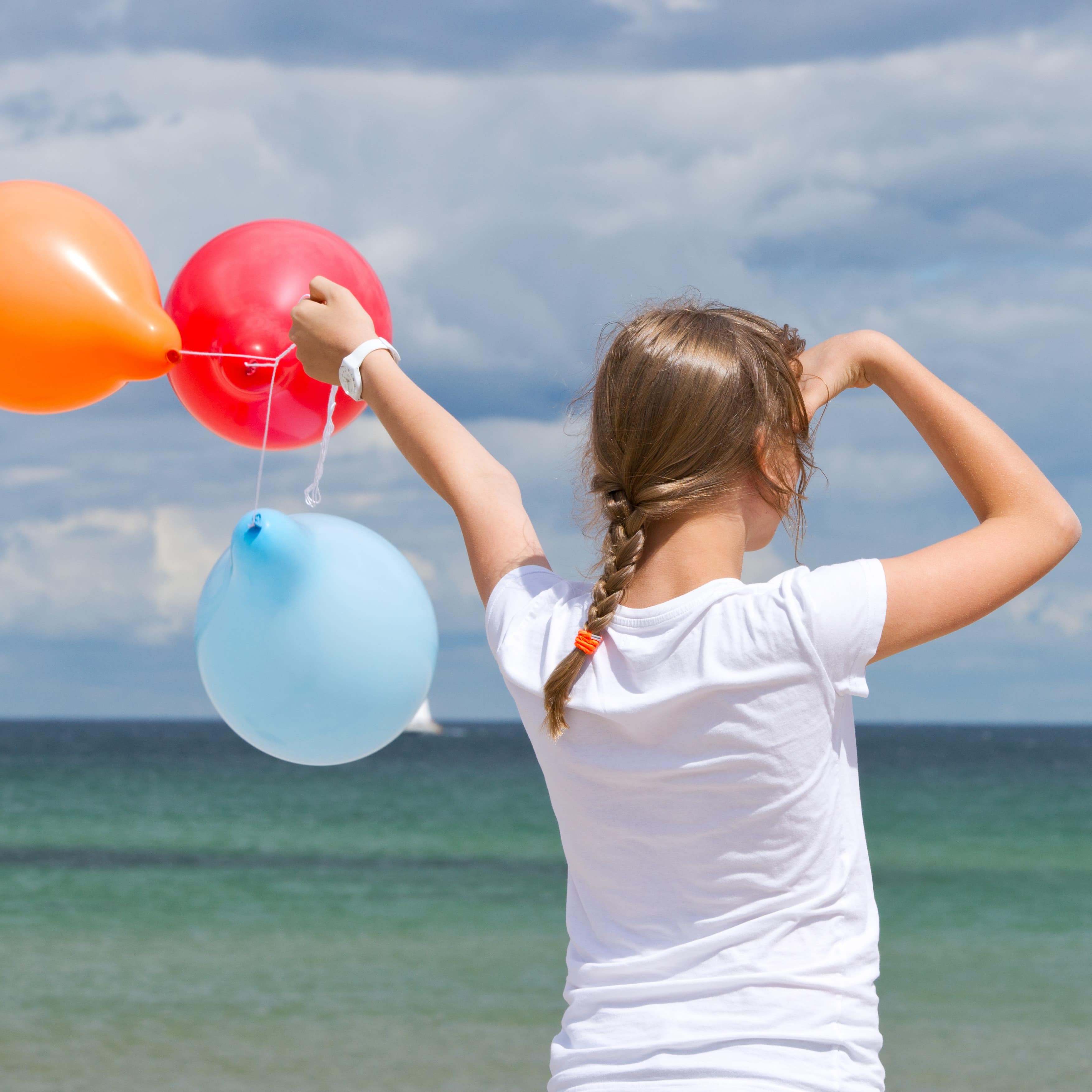 Rückenansicht: Ein Mädchen mit geflochtenem Zopf und 3 bunten Luftballons blickt auf des Meer.