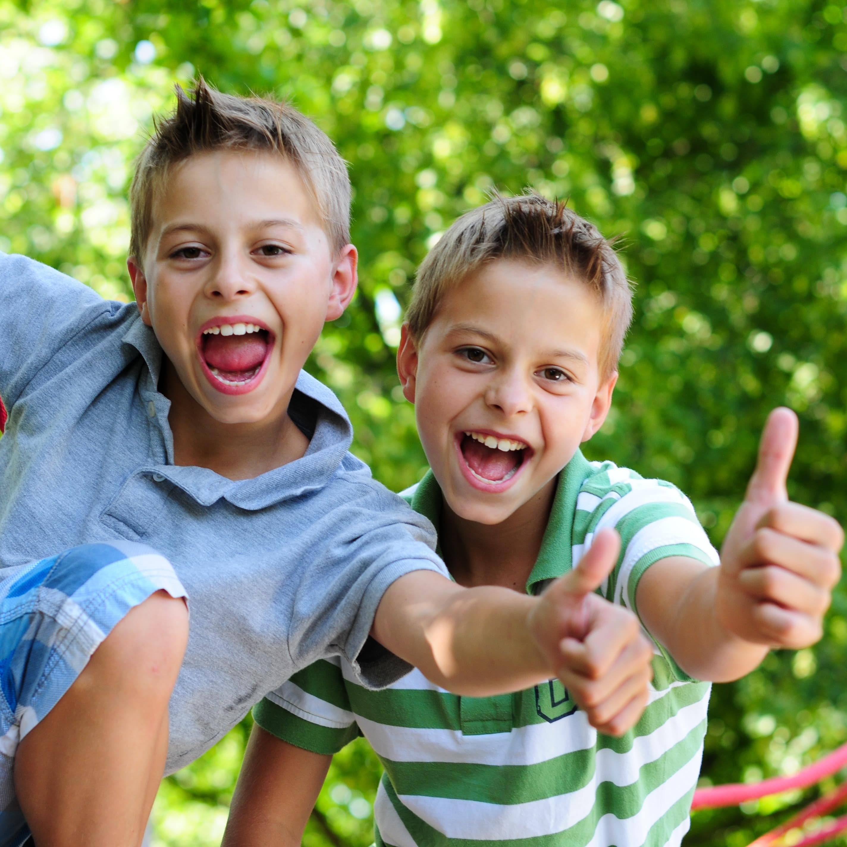 2 Jungen in Sommerkleidung auf einem Abenteuerspielplatz lachen in die Kamera und geben ein „Thumbs-up“.