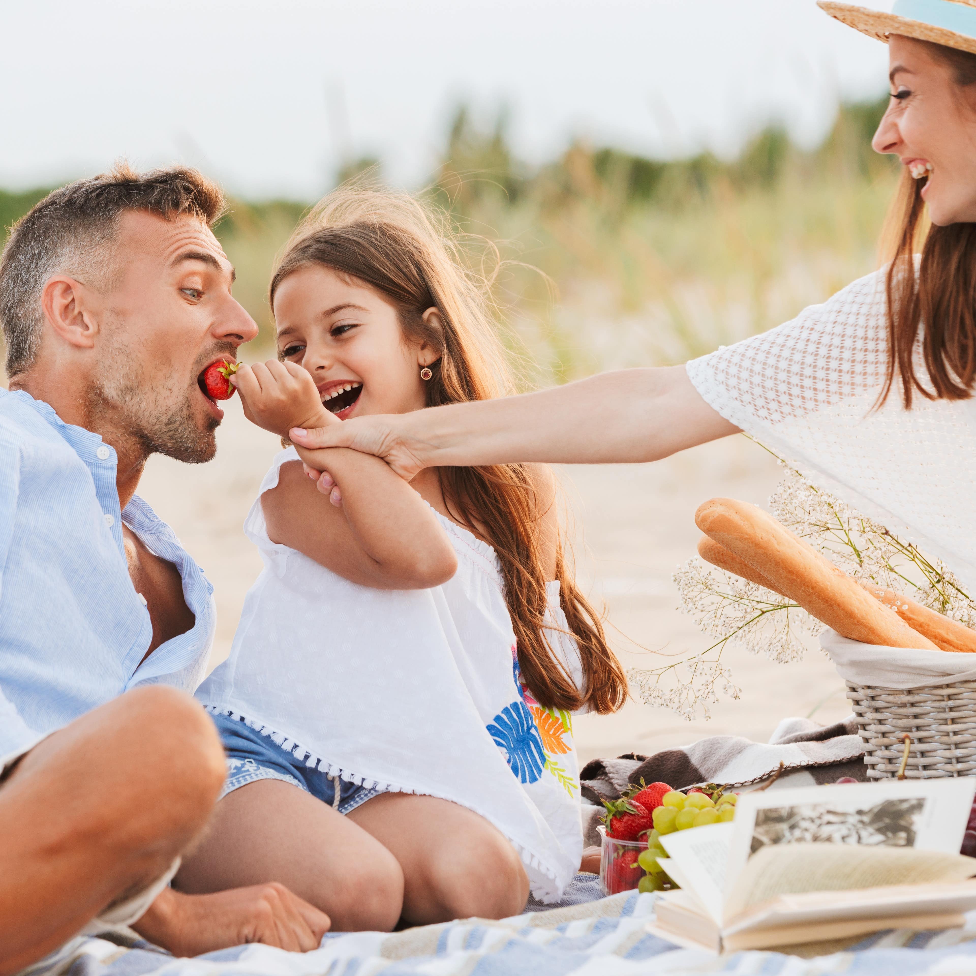 Picknick am Strand: Mutter und Tochter füttern den Vater mit einer Erdbeere.