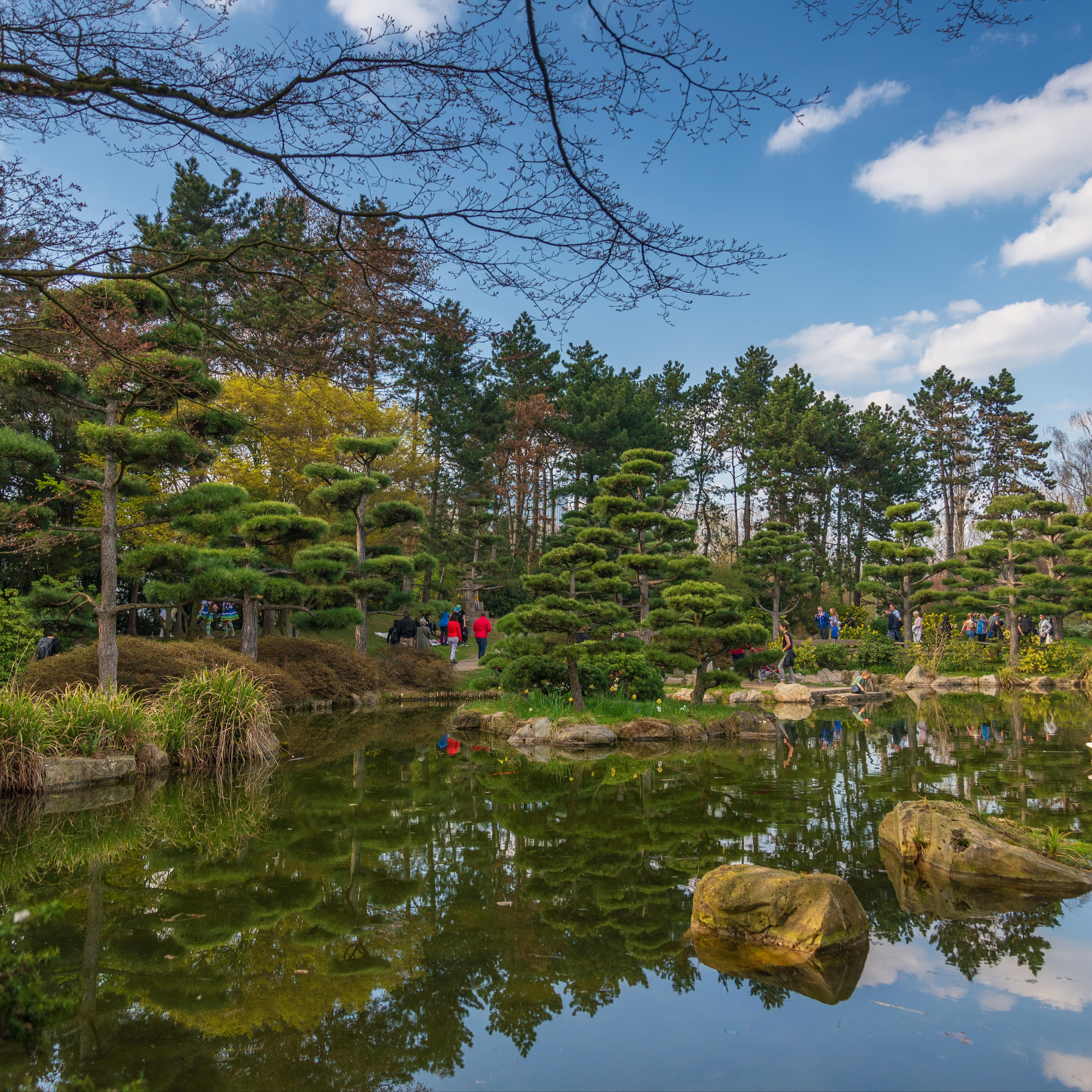 Blick über einen Teich und Bäume im Japanischen Garten, überall spazieren Leute.