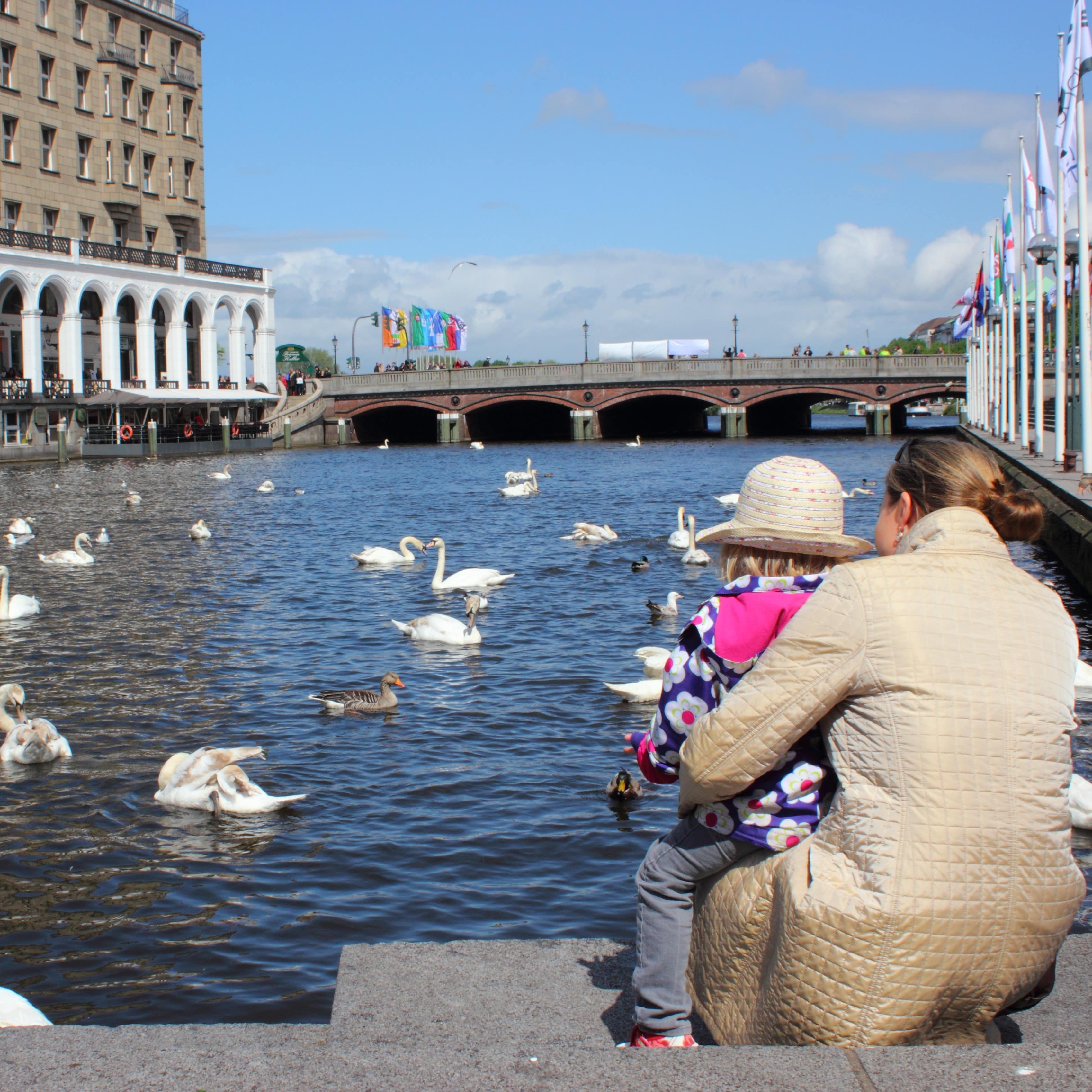 Mutter und Tochter füttern am Alsterufer Enten und Schwäne. Die Sonne scheint.