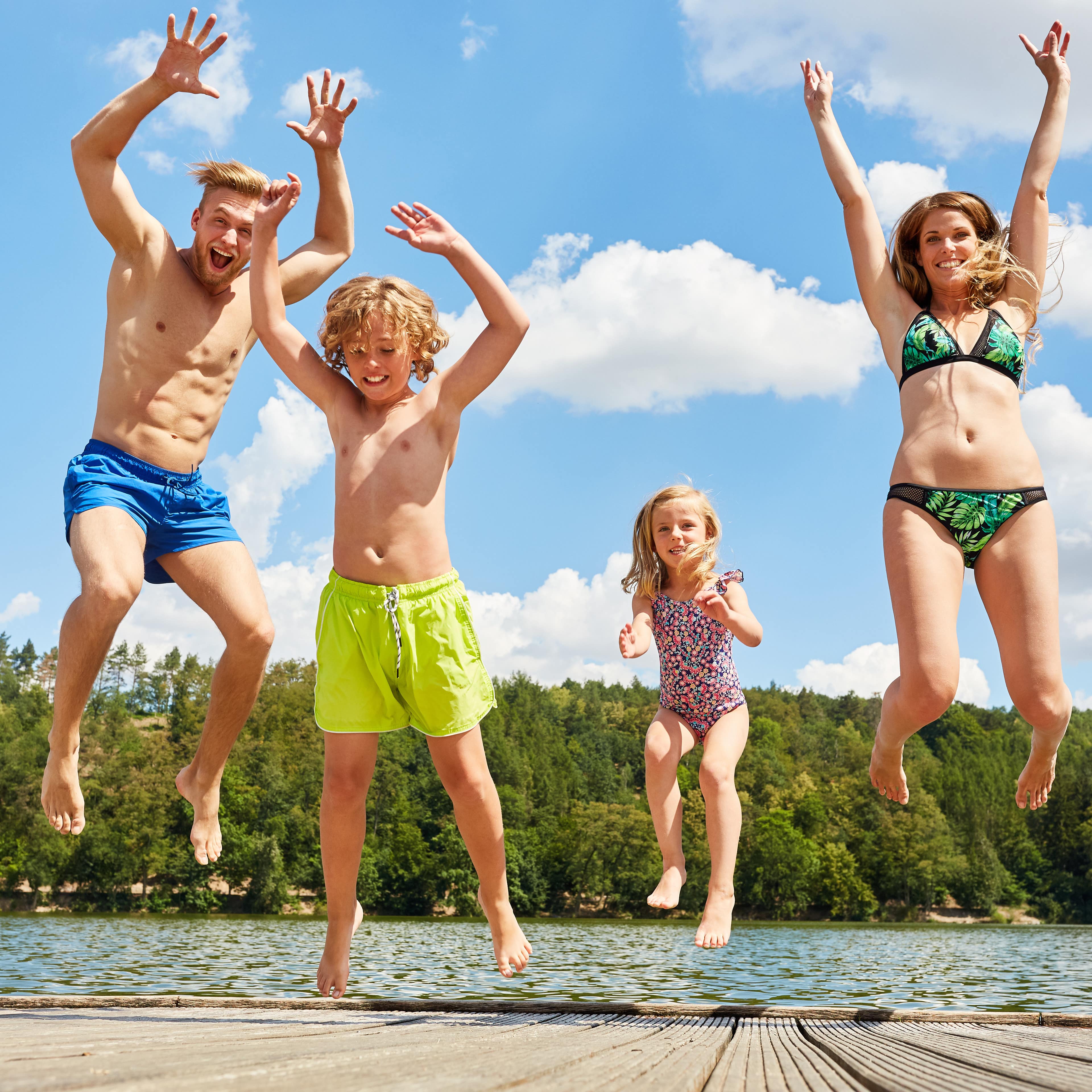 Familie mit 2 Kindern in Badekleidung springt auf einem Badesteg in die Luft.