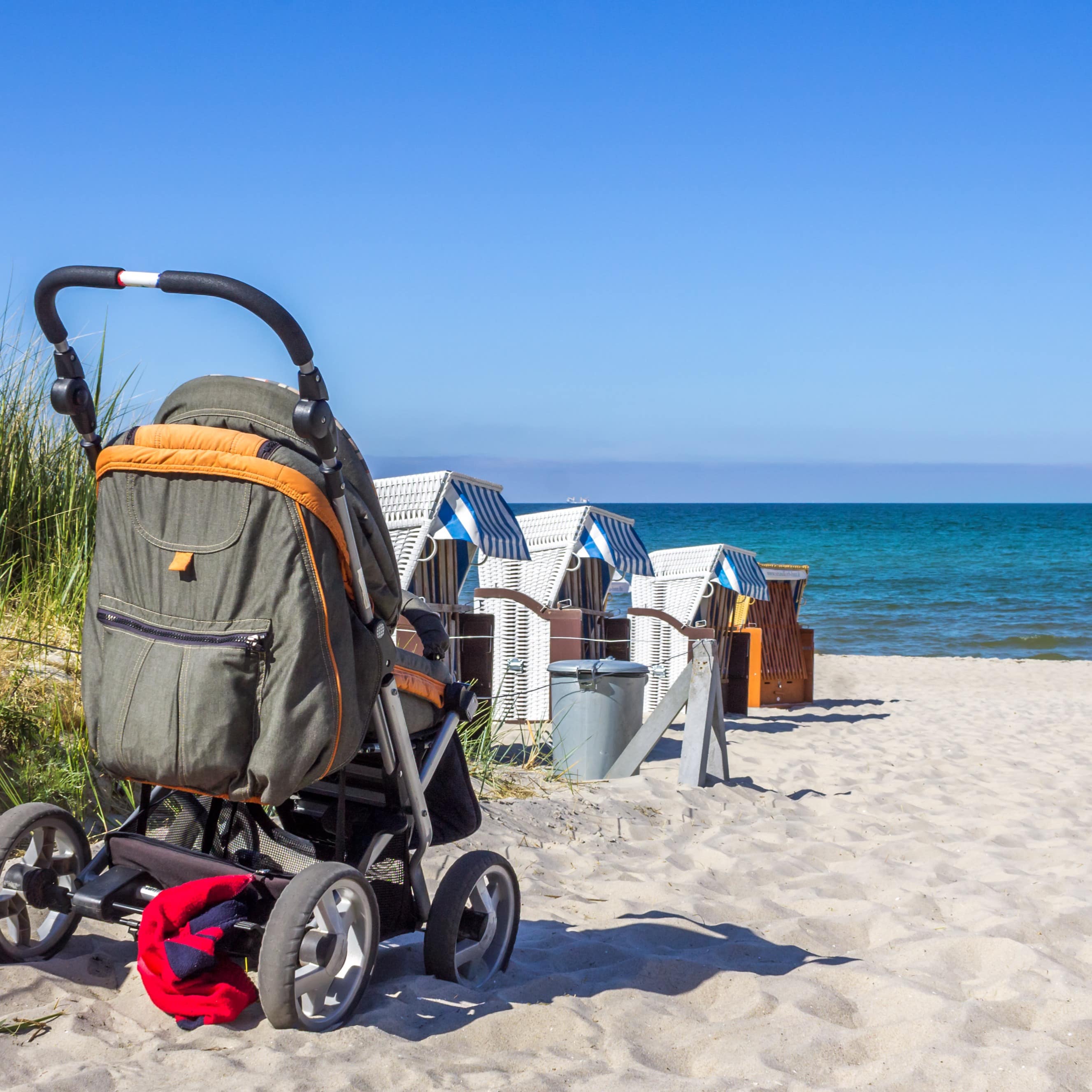 Ein Kinderwagen parkt am Zugang zum Strand, weiter vorne Strandkörbe und das Meer