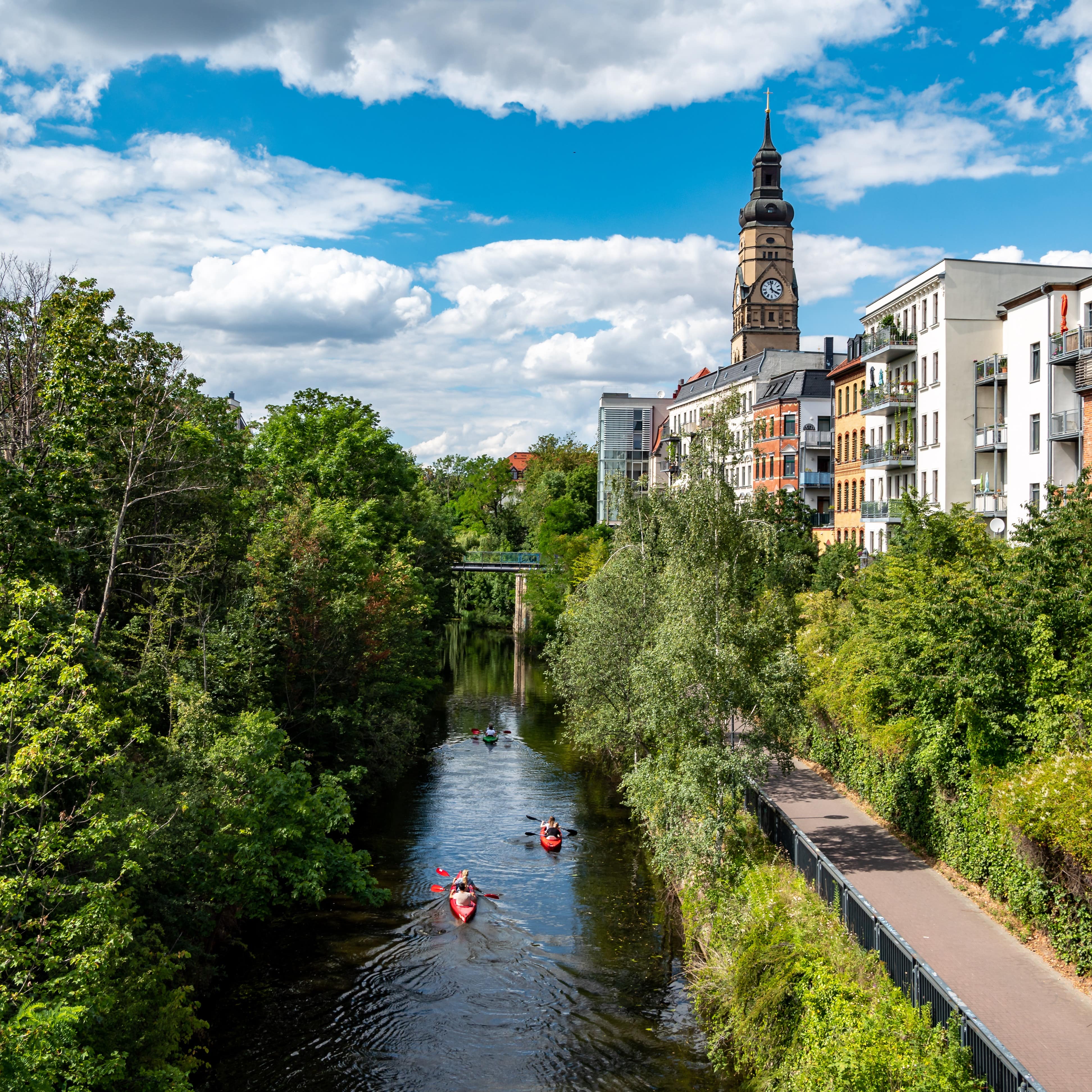 Blick von oben auf Personen in Kajaks auf dem Kanal, links Bäume, rechts ein Spazierweg und Häuser.