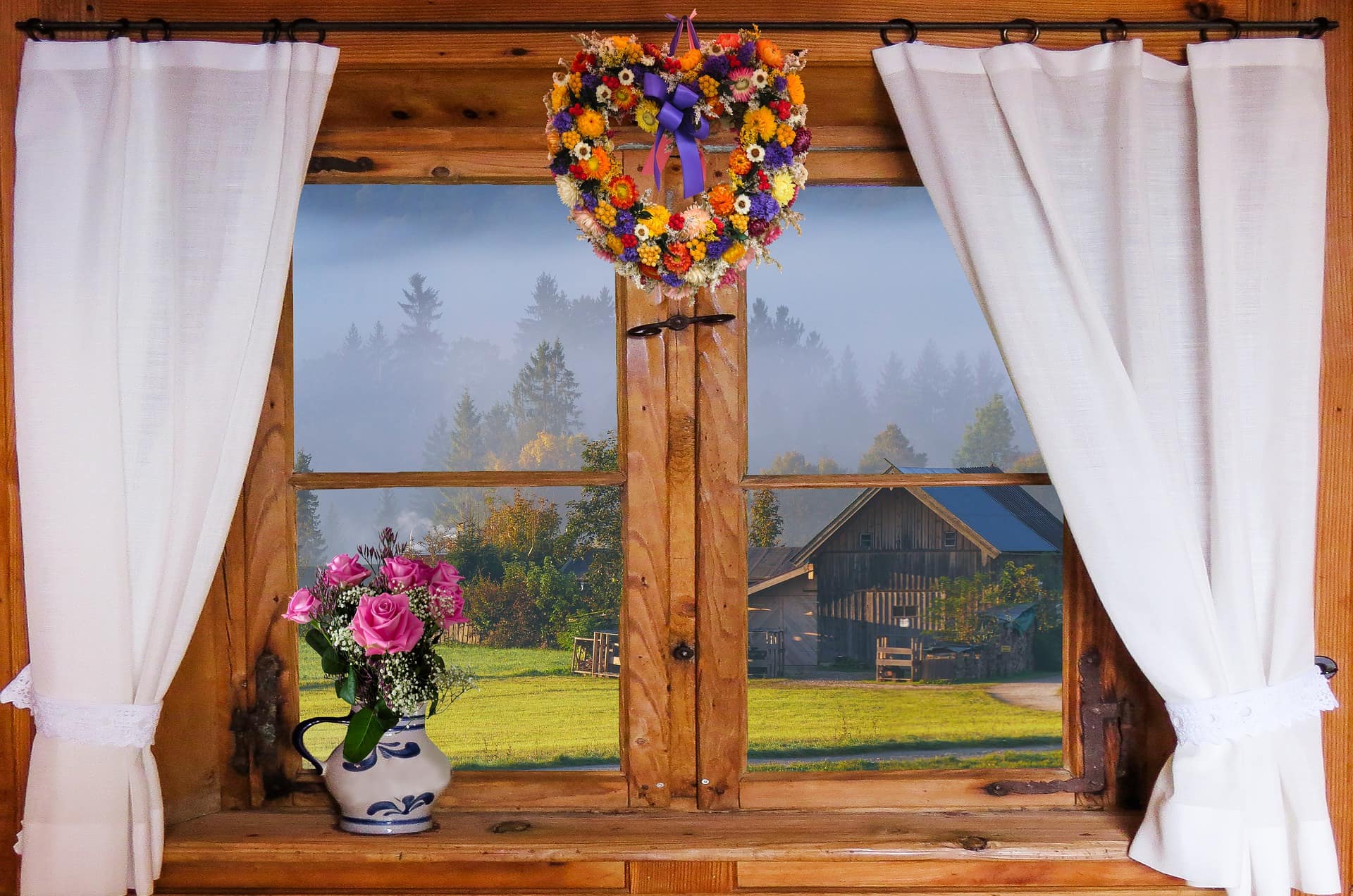 Blick aus dem Fenster mit weißen Gardinen und Blumenschmuck in einer Ferienwohnung in Bayern.