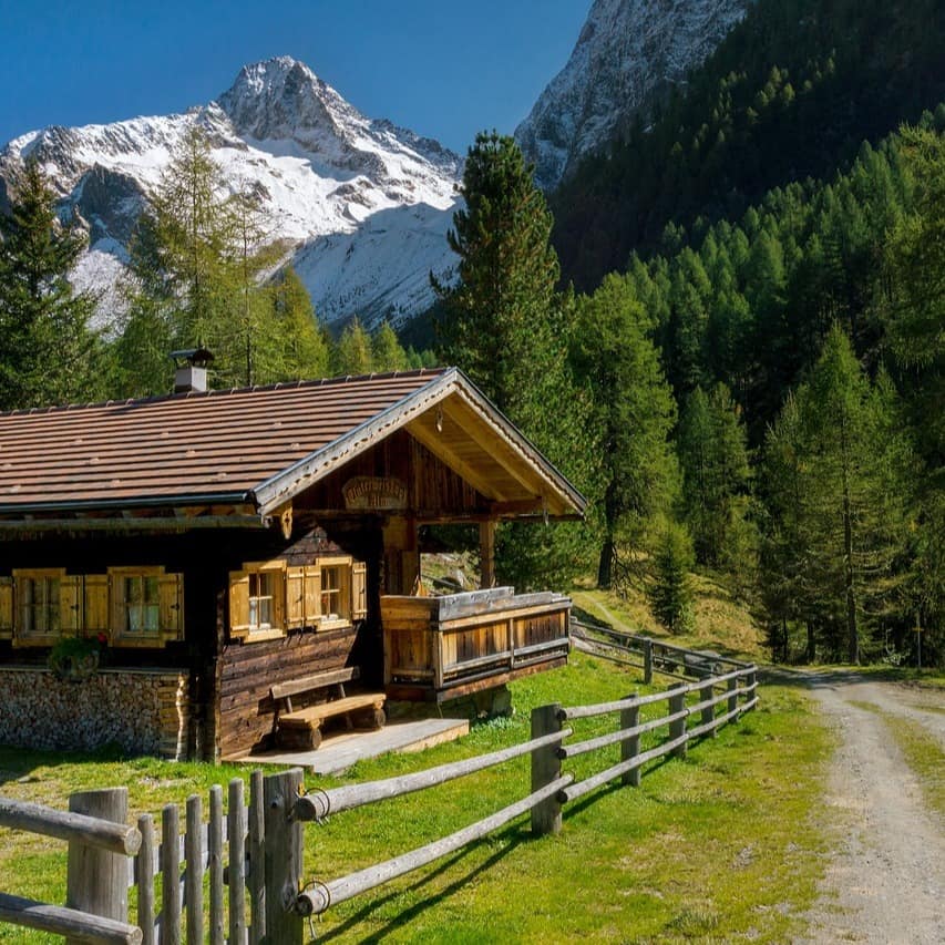 Eine eingezäunte Almhütte zwischen Wäldern und Bergen.